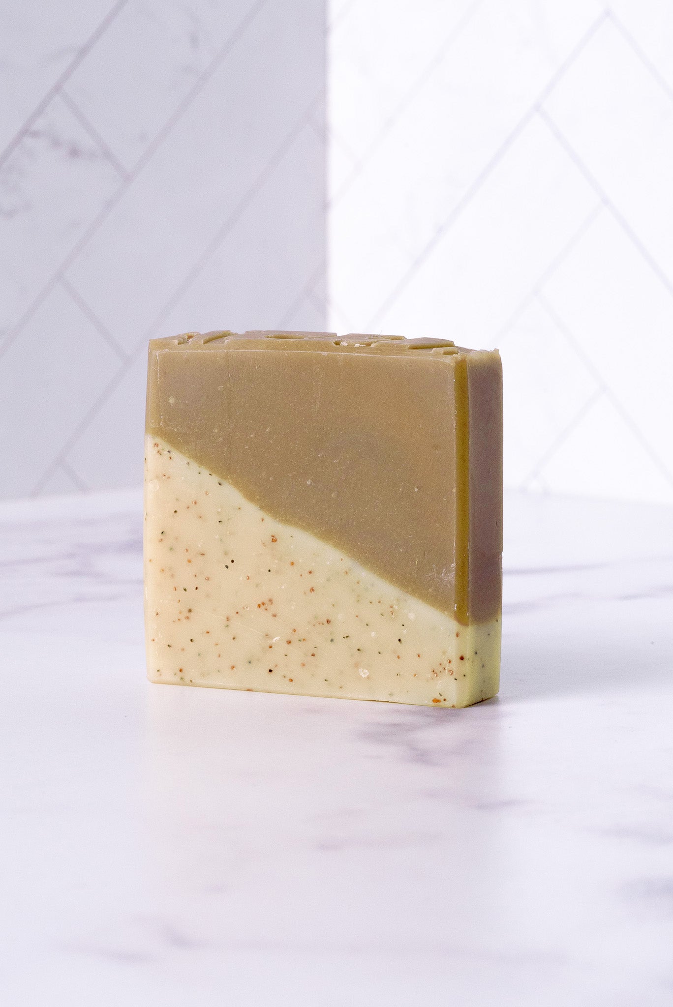 Rustic Strength’s Bar Soap | Sandalwood Patchouli is a rectangular cold process soap with a diagonal split—olive green over speckled cream—crafted with hydrating natural ingredients and shown on white marble against tiled background.