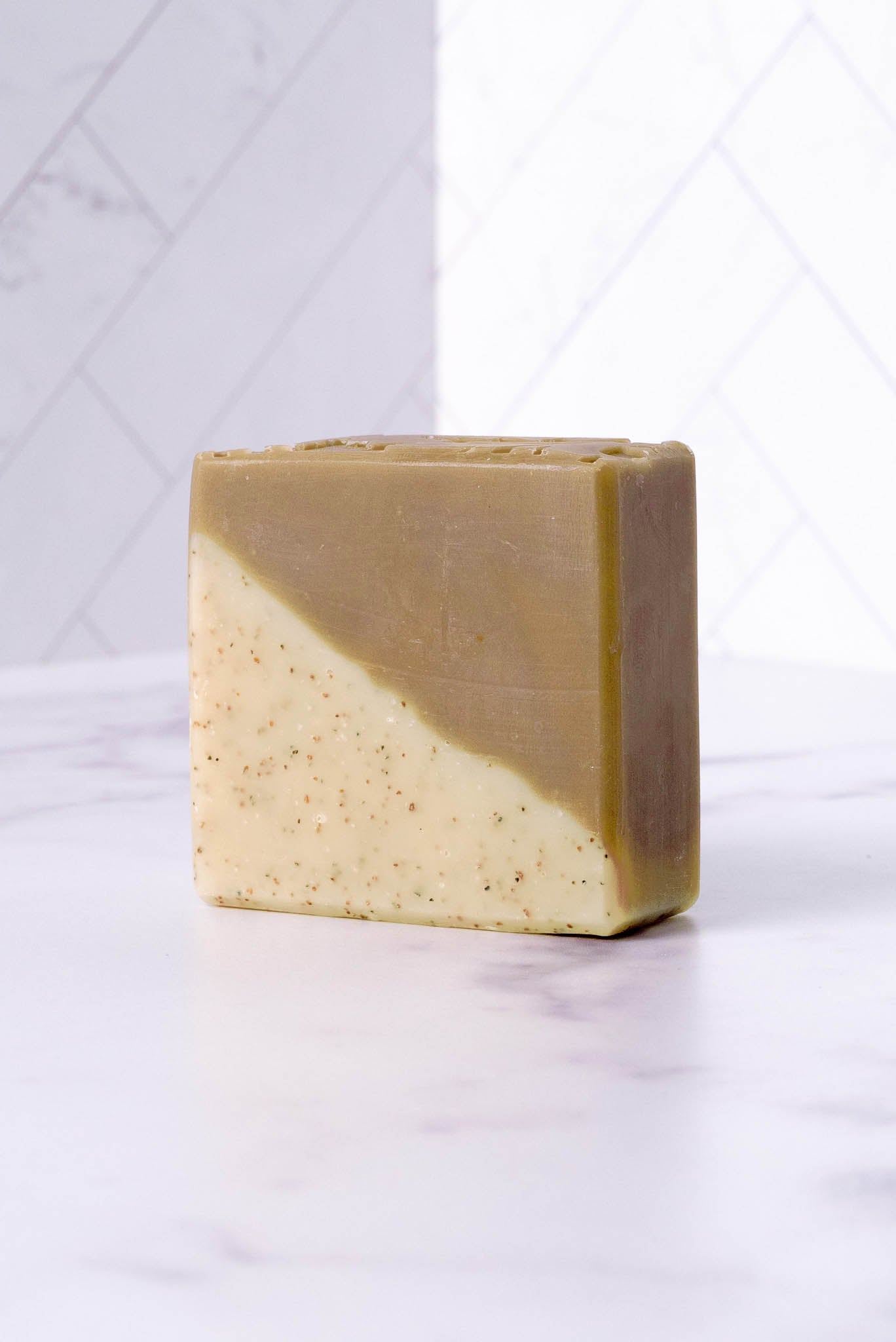 The Rustic Strength Bar Soap | Sandalwood Patchouli features two diagonal layers—one tan with exfoliating specks, the other solid light brown—and rests on a white marble surface against a tiled background.