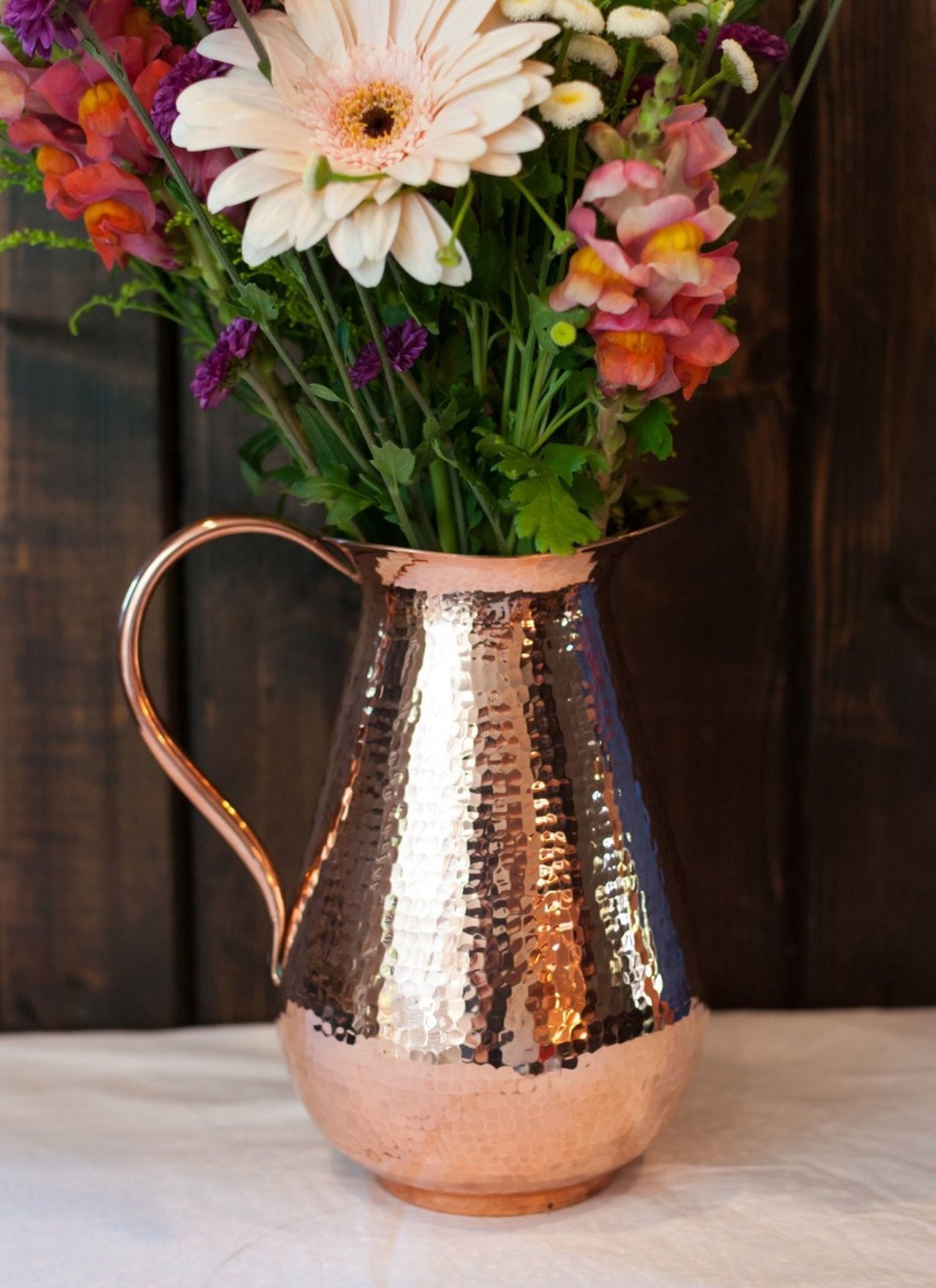 Sertodo Bisotun copper water pitcher with a copper handle, shown in a lifestyle setting on a wooden surface.