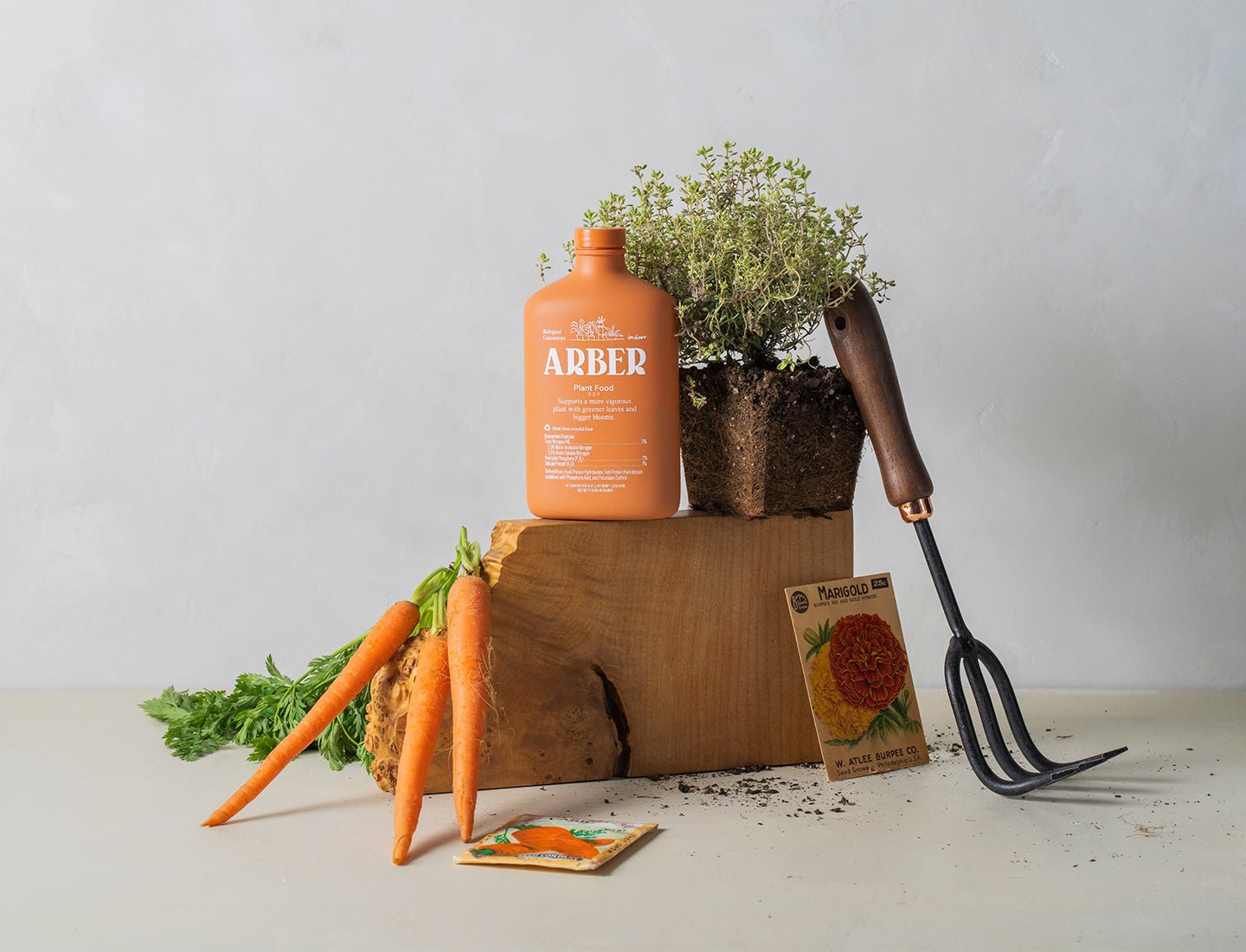 A gardening scene showcases fresh carrots, a wooden block, Arber Organic Plant Food, a potted herb, a hand cultivator, and seed packets—displaying indoor gardening essentials against a plain light background.