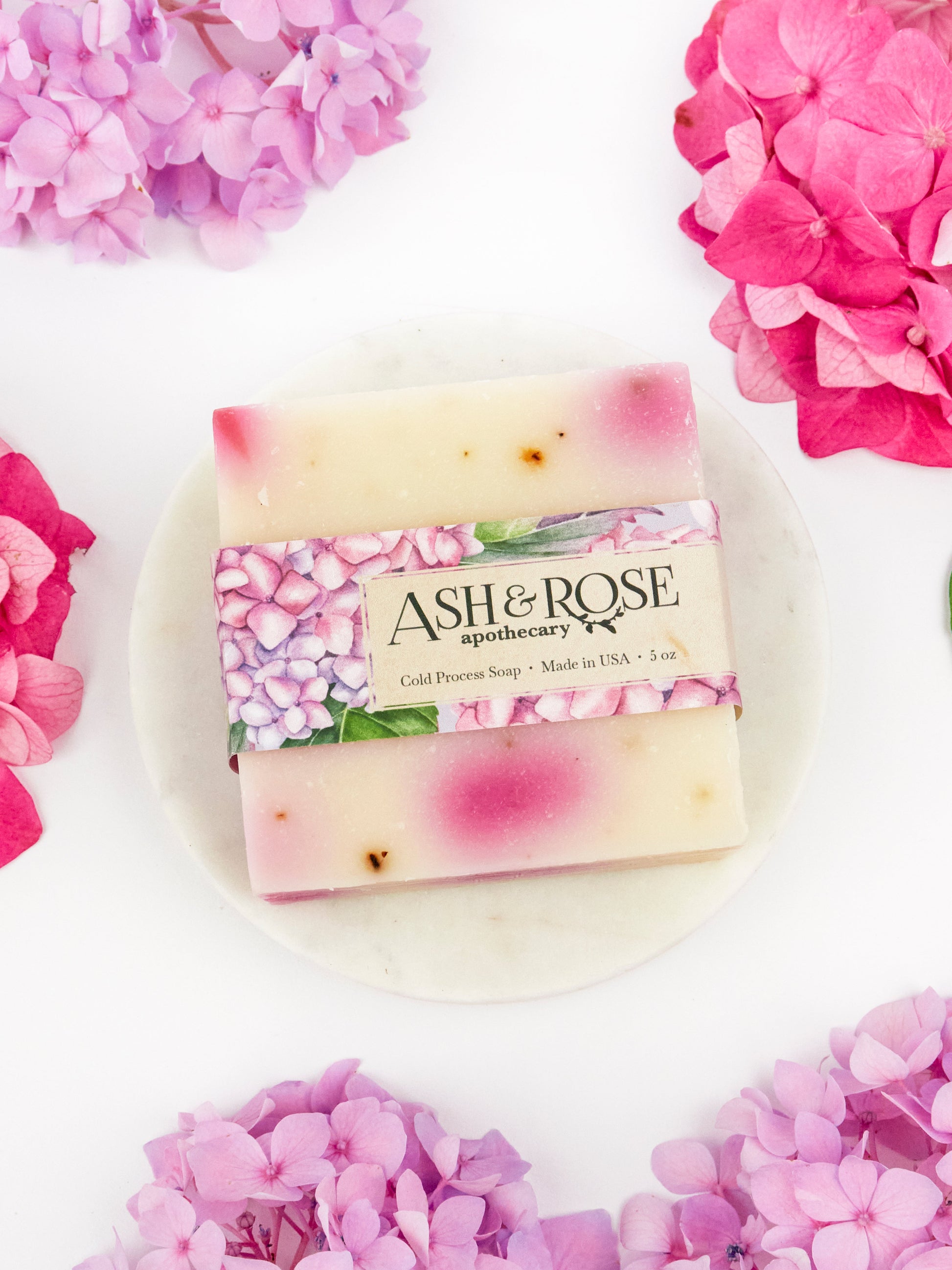 The Ash & Rose Pink Hydrangea Soap Scrub Bar, with its pink and white marbled design, sits on a round white dish among pink hydrangea flowers. Its label displays a floral motif and product details.
