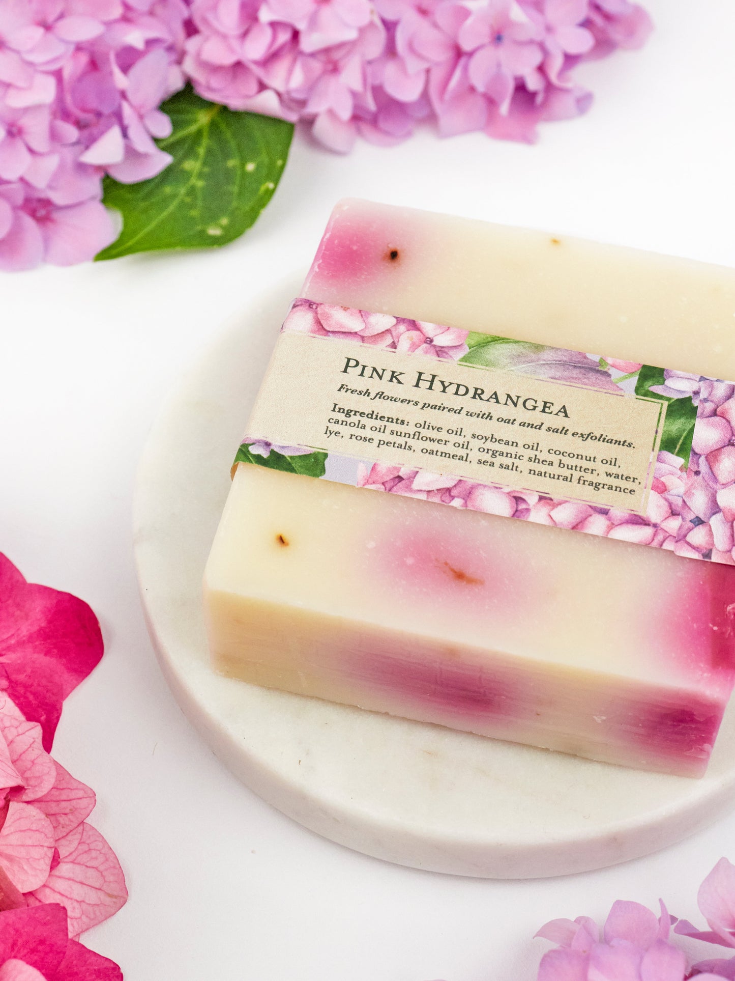 A bar of Ash & Rose’s Pink Hydrangea Soap Scrub Bar, in purple and cream hues, rests on a white marble dish surrounded by pink and purple hydrangeas. The label features floral designs and lists the ingredients.