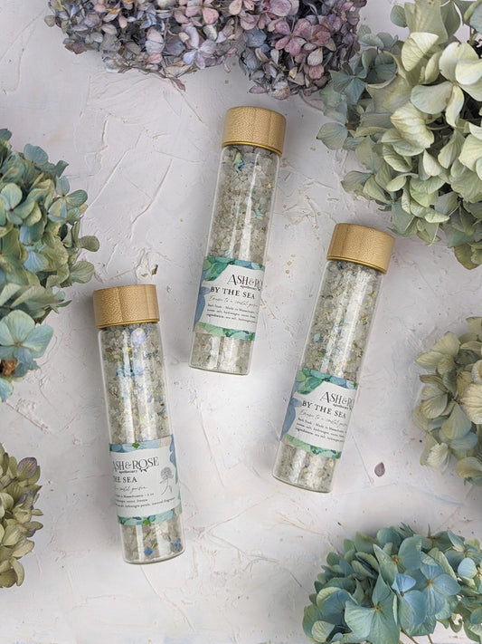 Three BY THE SEA Bath Soak Vials by Ash & Rose are arranged on a light textured surface, surrounded by clusters of pastel hydrangea flowers.