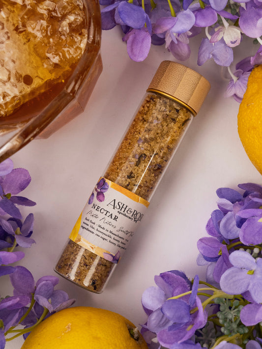 A glass vial labeled "NECTAR Bath Soak" by Ash & Rose, filled with brown granules and surrounded by lilac petals, lemons, and a bowl of honey on a white surface, highlighting its natural essence.