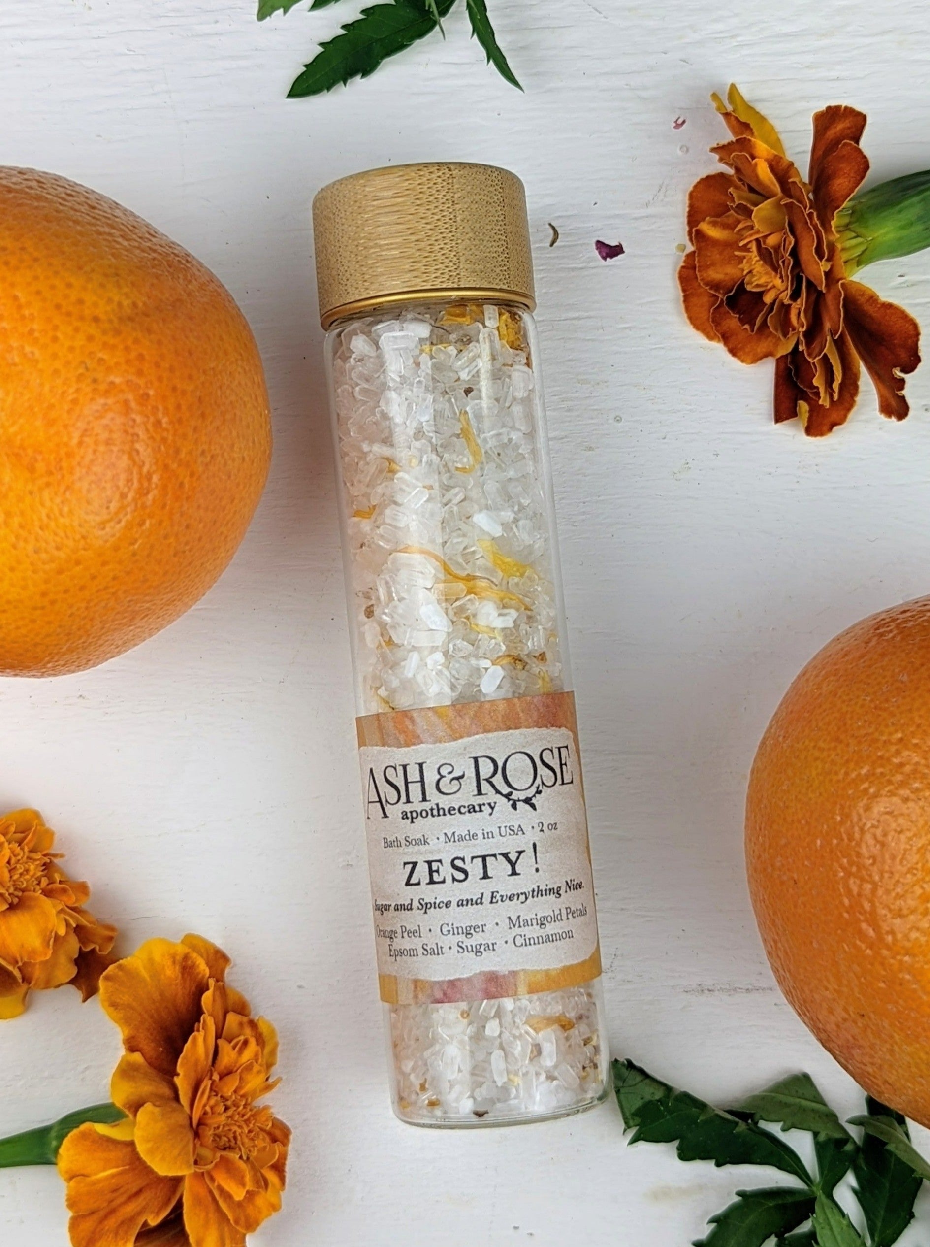 A clear vial of Ash & Rose ZESTY! Orange Ginger Bath Soak is surrounded by marigold flowers and oranges, showcasing visible Epsom salt crystals and spices on a white background.