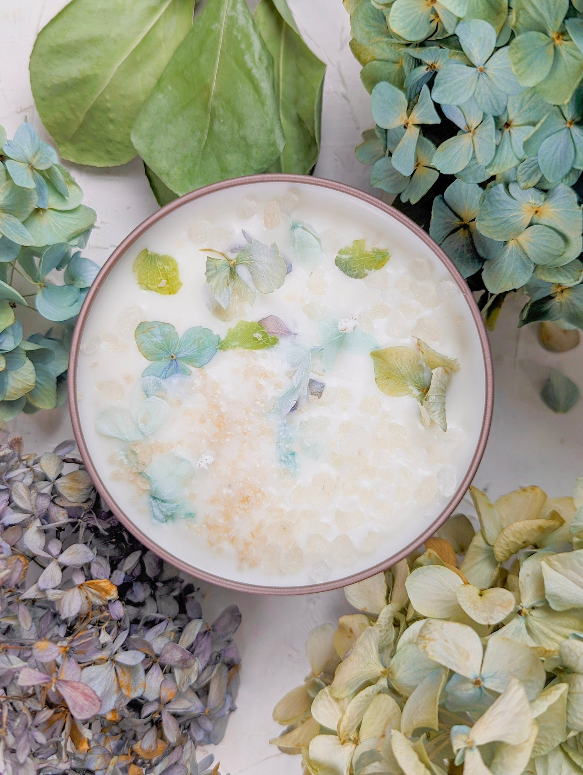 A creamy white dessert topped with pastel edible petals, surrounded by hydrangeas, recalls the fresh elegance of Ash & Rose’s BY THE SEA Hydrangea Sea Salt Candle on a light background.