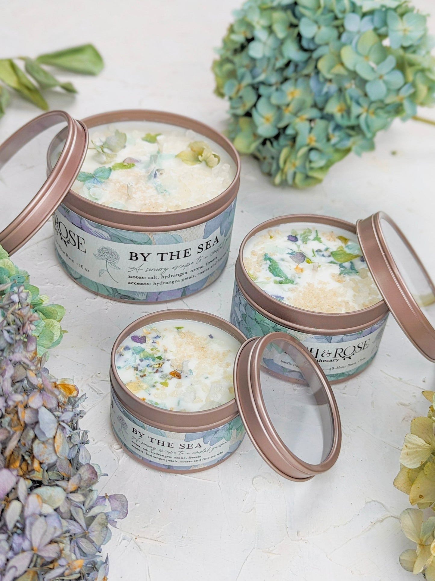 Three round tins labeled BY THE SEA by Ash & Rose, including a Hydrangea Sea Salt Candle, are filled with decorative bath products and displayed beside green and purple hydrangeas. The tins feature rose-gold lids, two shown open.