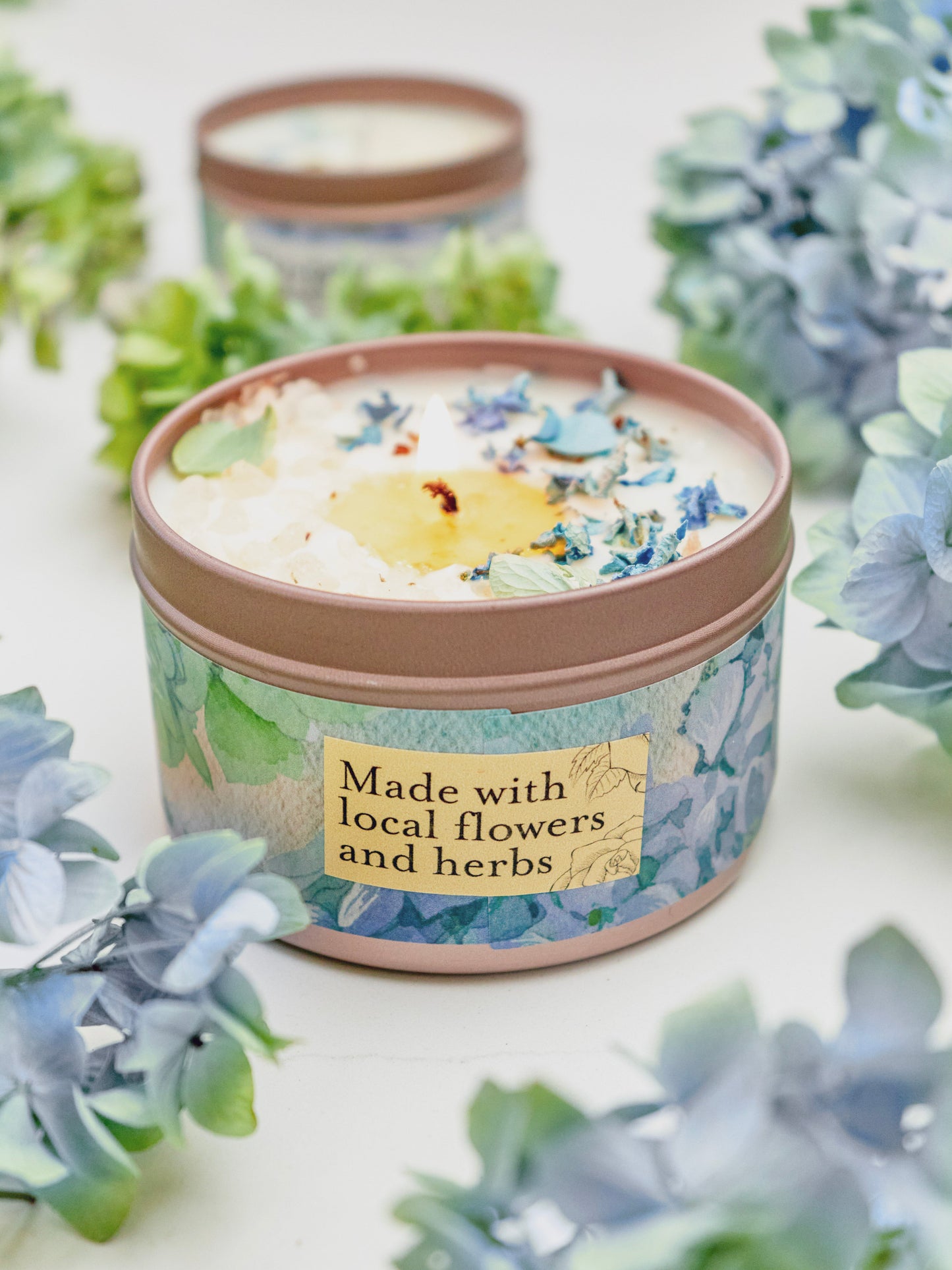The Ash & Rose BY THE SEA Hydrangea Sea Salt Candle sits among light blue and green hydrangeas, its label reading Made with local flowers and herbs, evoking a fresh coastal garden vibe. Another similar soy wax candle tin is blurred in the background.