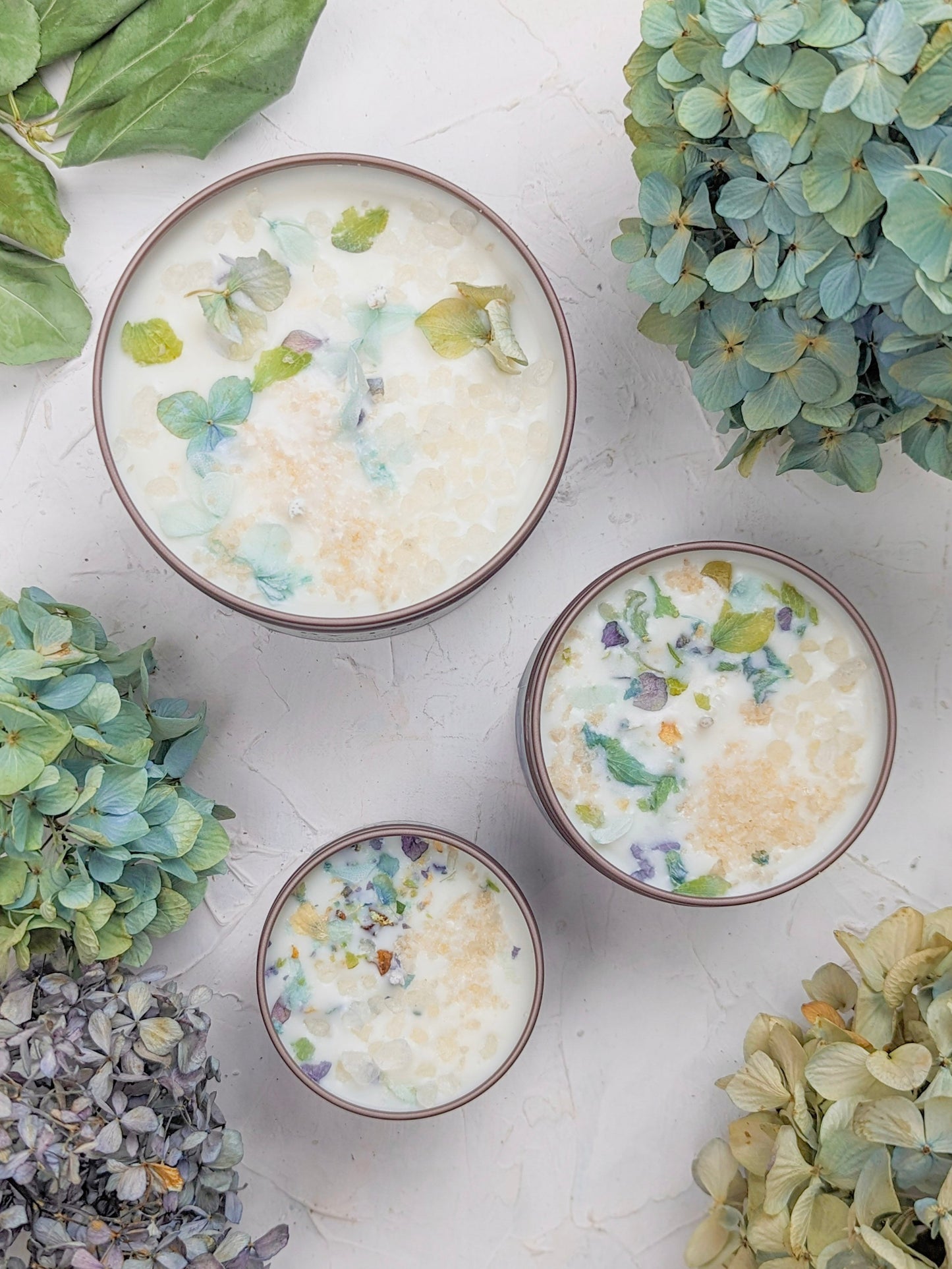 Three bowls of creamy dessert, topped with edible flower petals, sit on a textured surface surrounded by green and blue hydrangeas and the softly glowing "BY THE SEA Hydrangea Sea Salt Candle" from Ash & Rose.