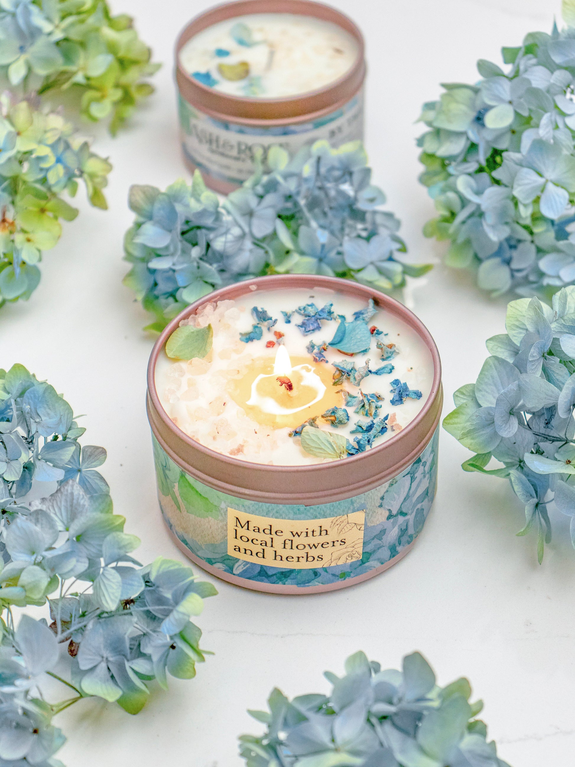 The BY THE SEA Hydrangea Sea Salt Candle by Ash & Rose glows in a decorated tin labeled "Made with local flowers and herbs," surrounded by blue hydrangea blooms, with another soy wax candle in the background on a white surface.