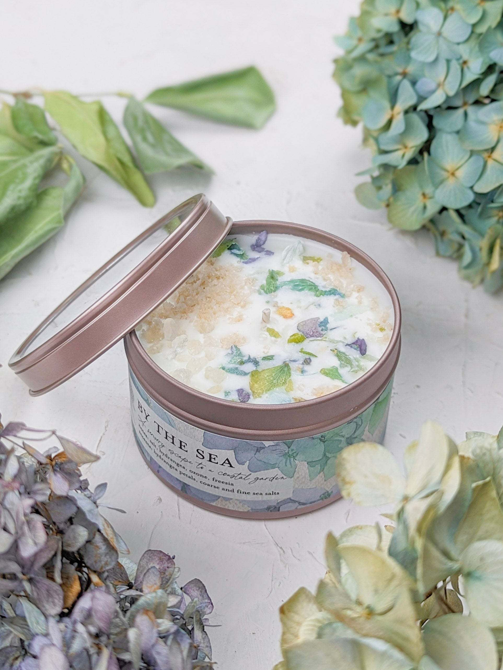 The BY THE SEA Hydrangea Sea Salt Candle by Ash & Rose is a soy wax candle in a round tin with a floral label, topped with colorful dried flowers and crystals, displayed open on white with dried hydrangeas and green leaves.