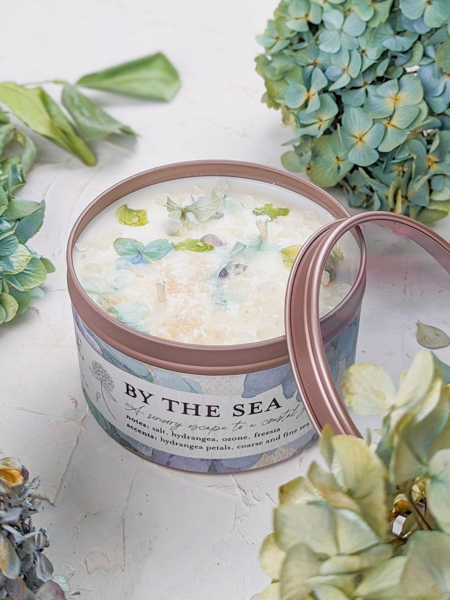 The BY THE SEA Hydrangea Sea Salt Candle by Ash & Rose is a round candle adorned with blue and green hydrangea petals and crystals, labeled on the surface, with its lid resting open beside this coastal-inspired floral candle.