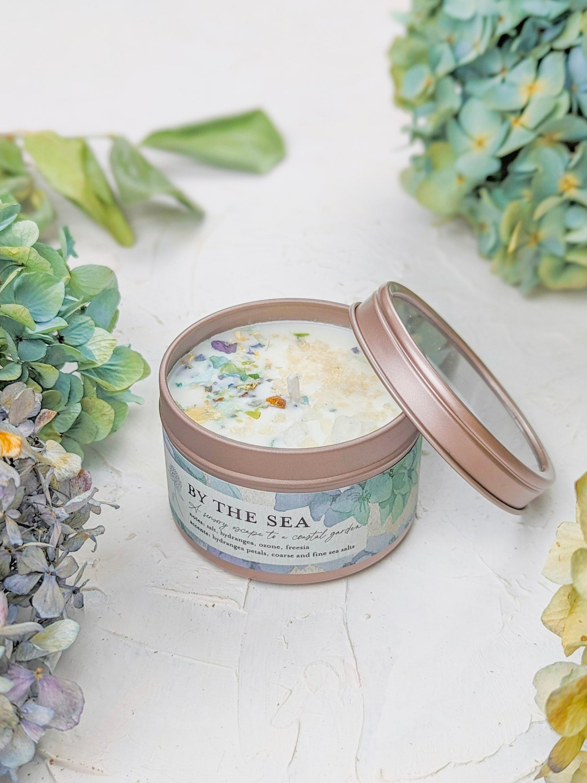 The BY THE SEA Hydrangea Sea Salt Candle by Ash & Rose, in a round tin with the lid off, sits on a white textured surface surrounded by green and blue hydrangeas. Dried flowers and crystals decorate the soy wax inside.