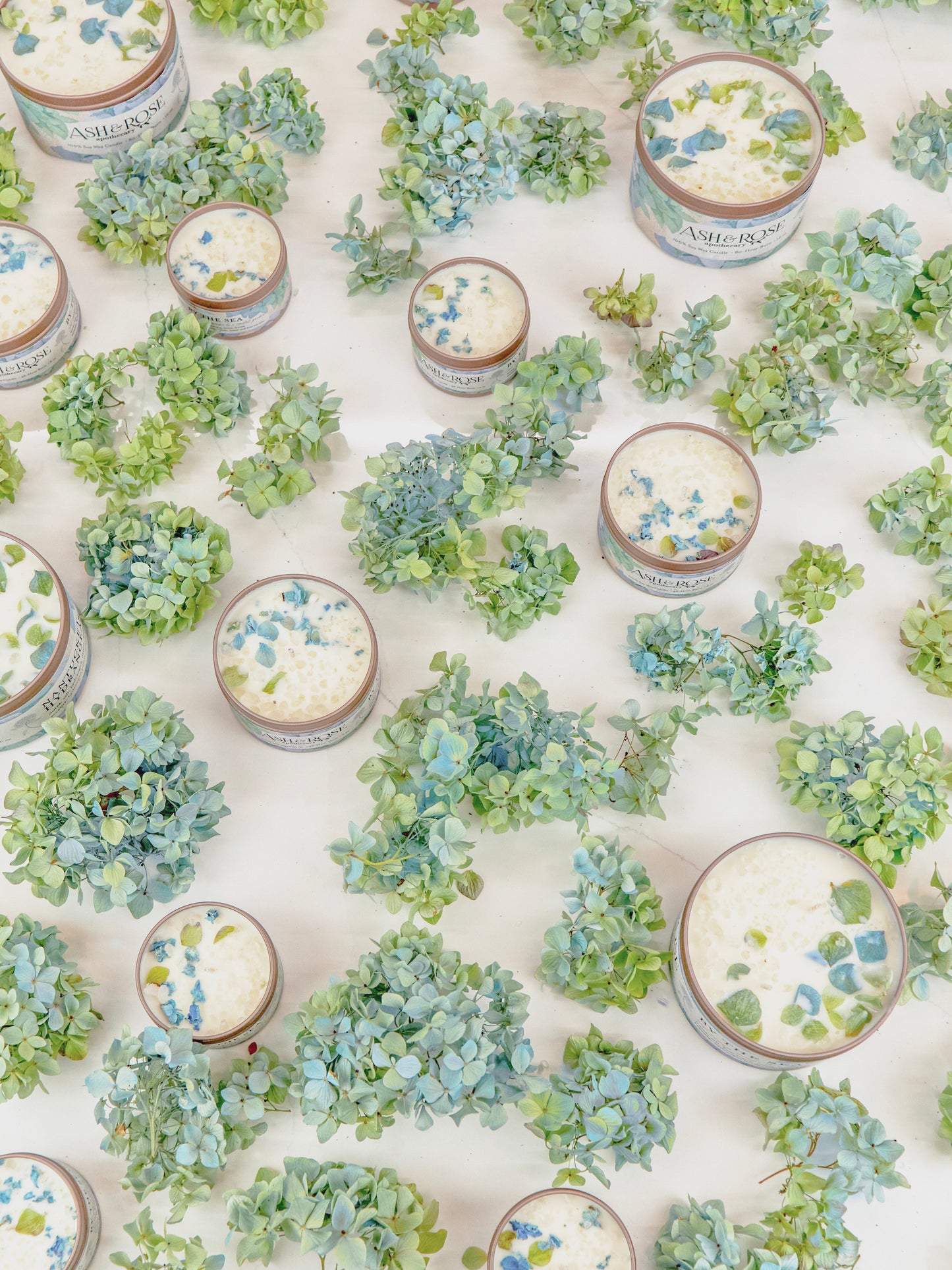 Several Ash & Rose BY THE SEA Hydrangea Sea Salt Candles, featuring blue and green hydrangea decorations, are neatly arranged on a white surface.