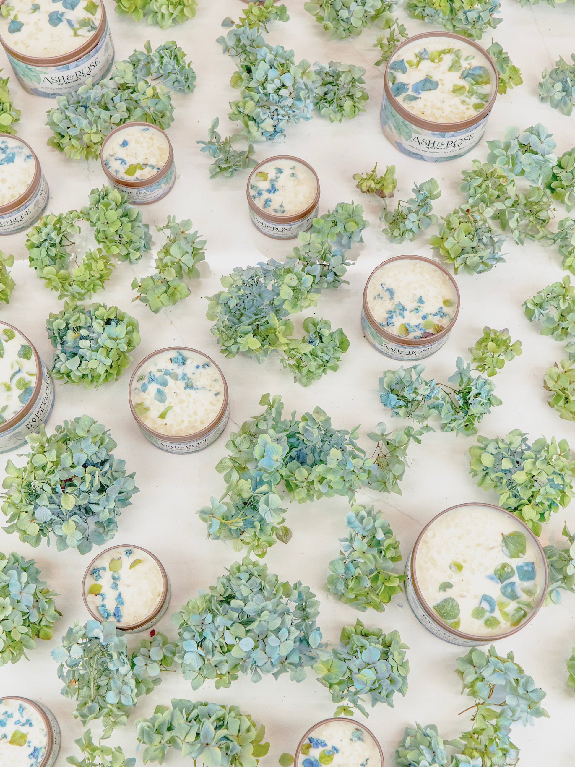 Several Ash & Rose BY THE SEA Hydrangea Sea Salt Candles, featuring blue and green hydrangea decorations, are neatly arranged on a white surface.