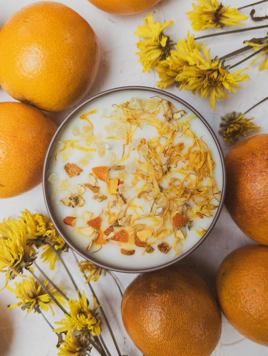 A bowl of yogurt with orange zest, dried petals, and granola sits beside the ZESTY! Orange Ginger Candle by Ash & Rose on a white surface, surrounded by fresh oranges and yellow flowers.