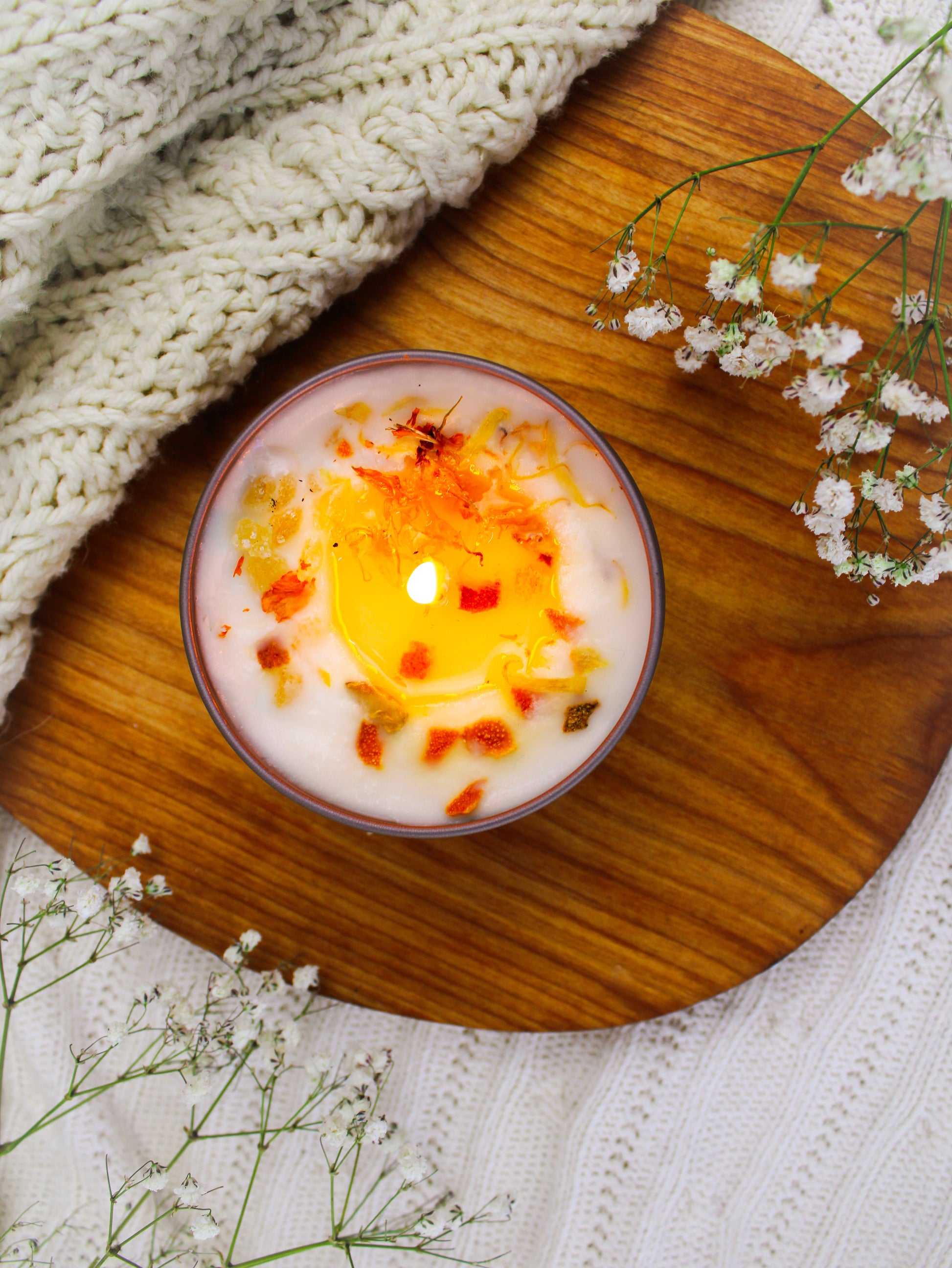 The ZESTY! Orange Ginger Candle by Ash & Rose, handmade and topped with dried flower petals and herbs, rests on a wooden tray beside a cream knitted blanket and small white flowers.