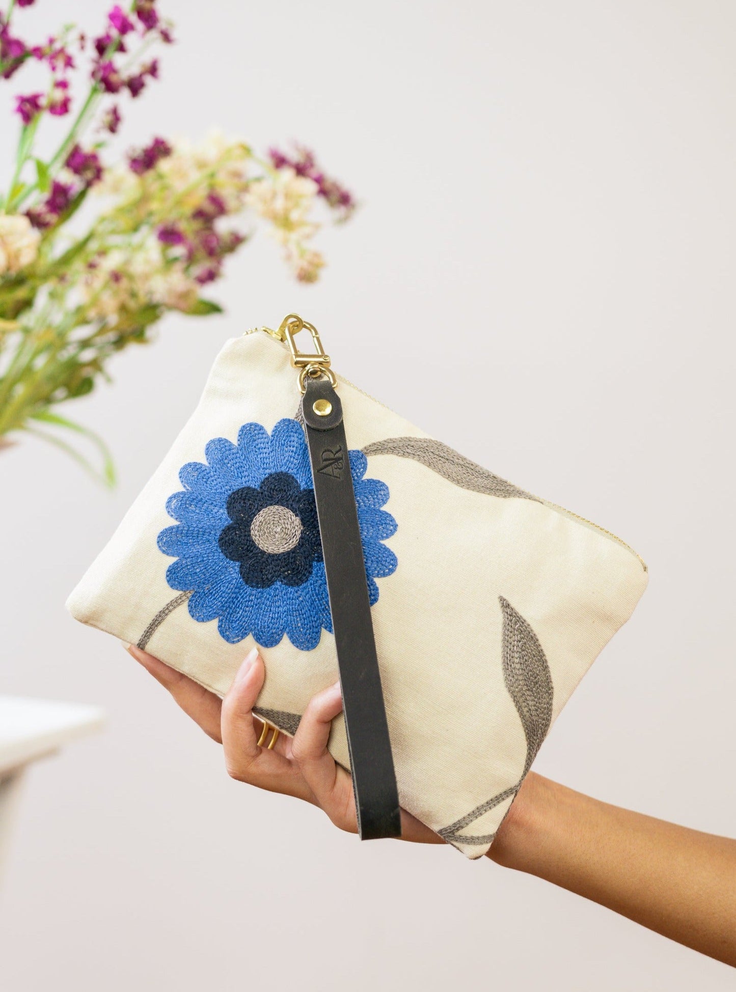 A hand holds the Blue Poppy Wristlet Purse by Ash & Rose—a cream linen designer bag with blue embroidered flower and black wrist strap. Blurred flowers in the background highlight this limited edition accessory.