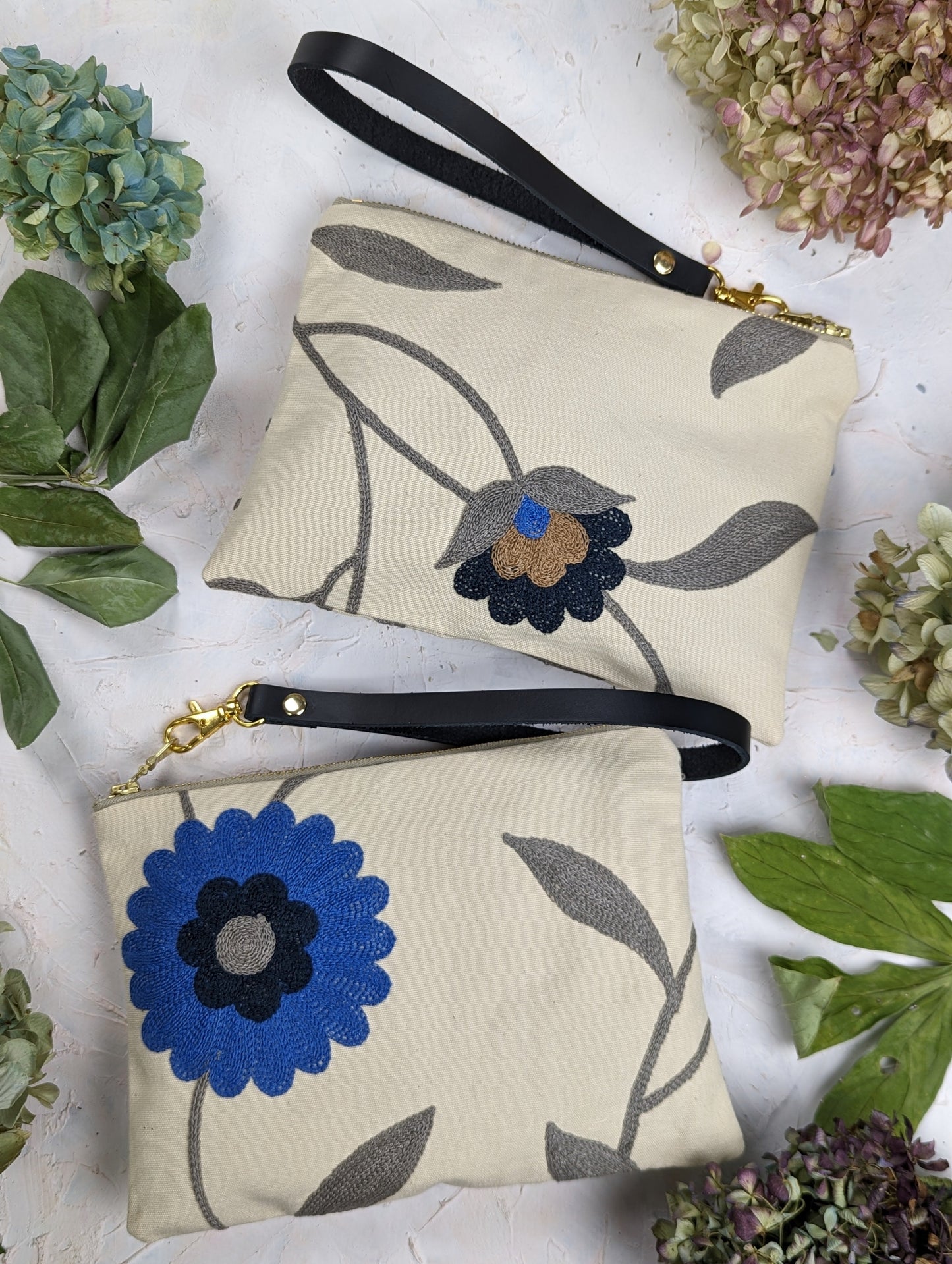 Two beige handmade wristlet pouches by Ash & Rose—a Blue Poppy Wristlet Purse with a bold blue flower and another with a black and brown bloom—are displayed amid green leaves and hydrangeas on a light surface.