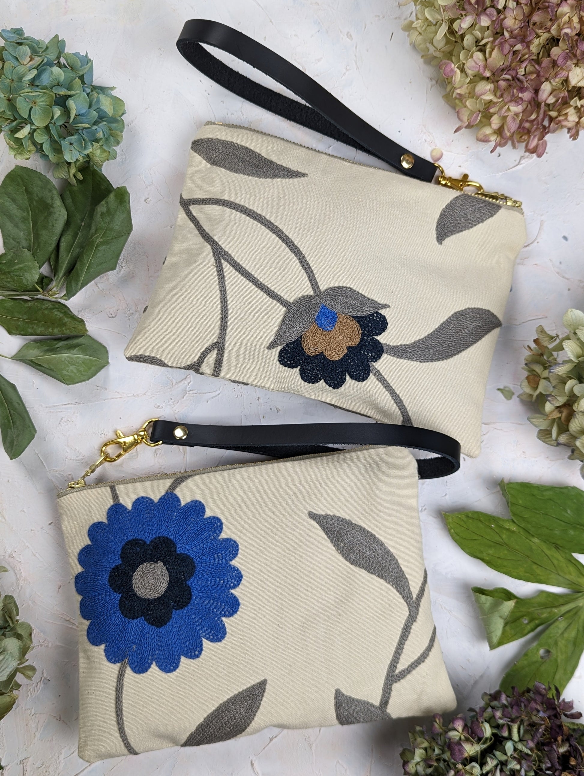 Two beige handmade wristlet pouches by Ash & Rose—a Blue Poppy Wristlet Purse with a bold blue flower and another with a black and brown bloom—are displayed amid green leaves and hydrangeas on a light surface.