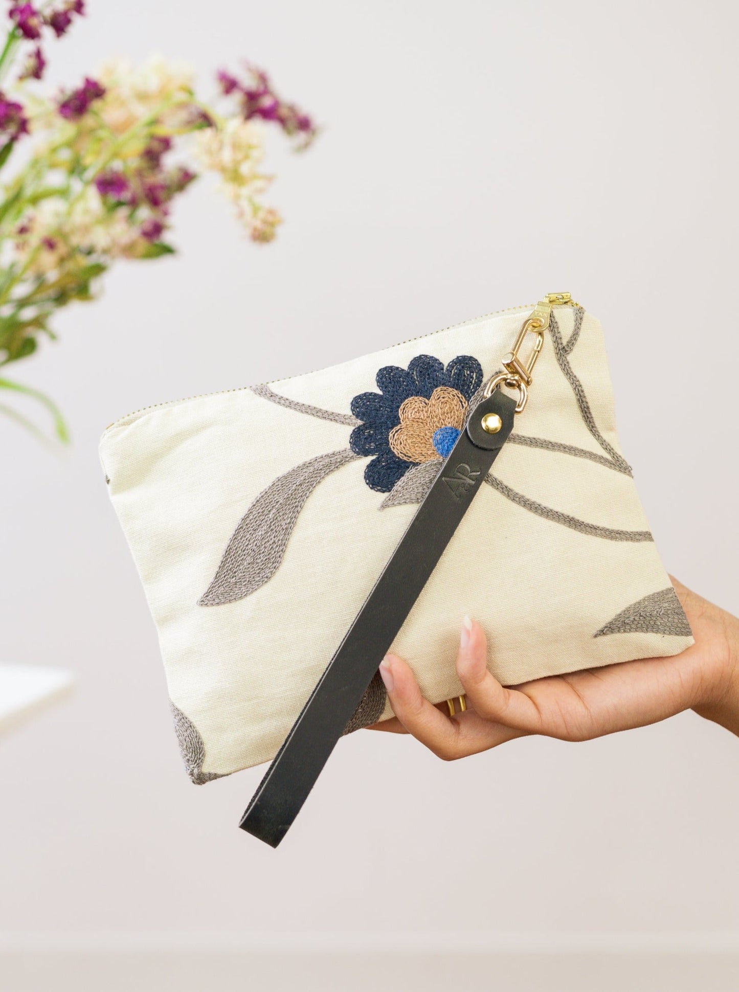 A hand holds the Ash & Rose Blue Poppy Wristlet Purse, a cream handmade pouch featuring a blue and brown floral design with a black strap; blurred flowers appear behind. This limited edition accessory enhances any ensemble.