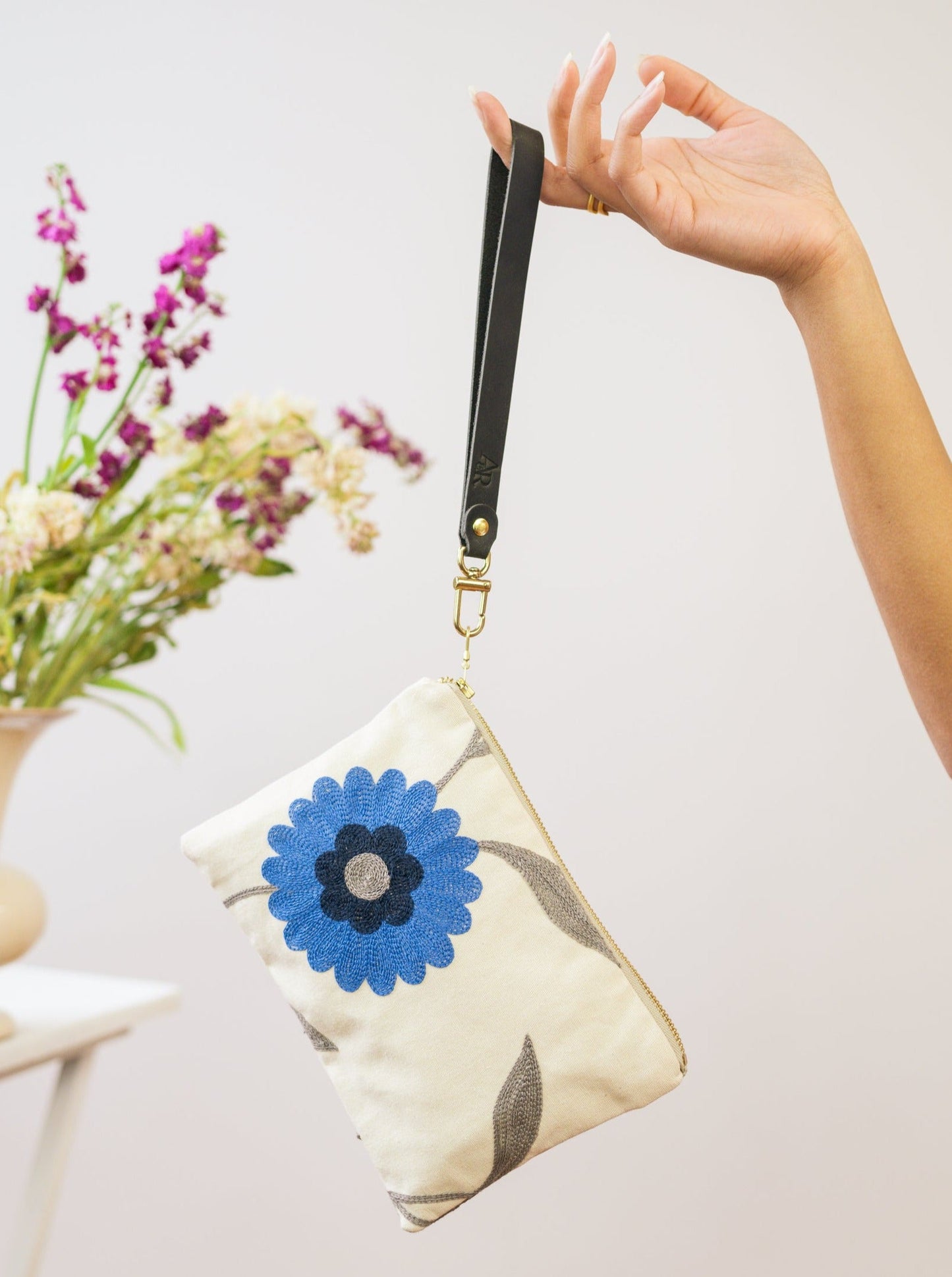 A hand holds the Ash & Rose Blue Poppy Wristlet Purse, a handmade bag featuring a black strap and blue flower design. In the background, a white vase with purple and white flowers enhances this limited edition accessory's charm.