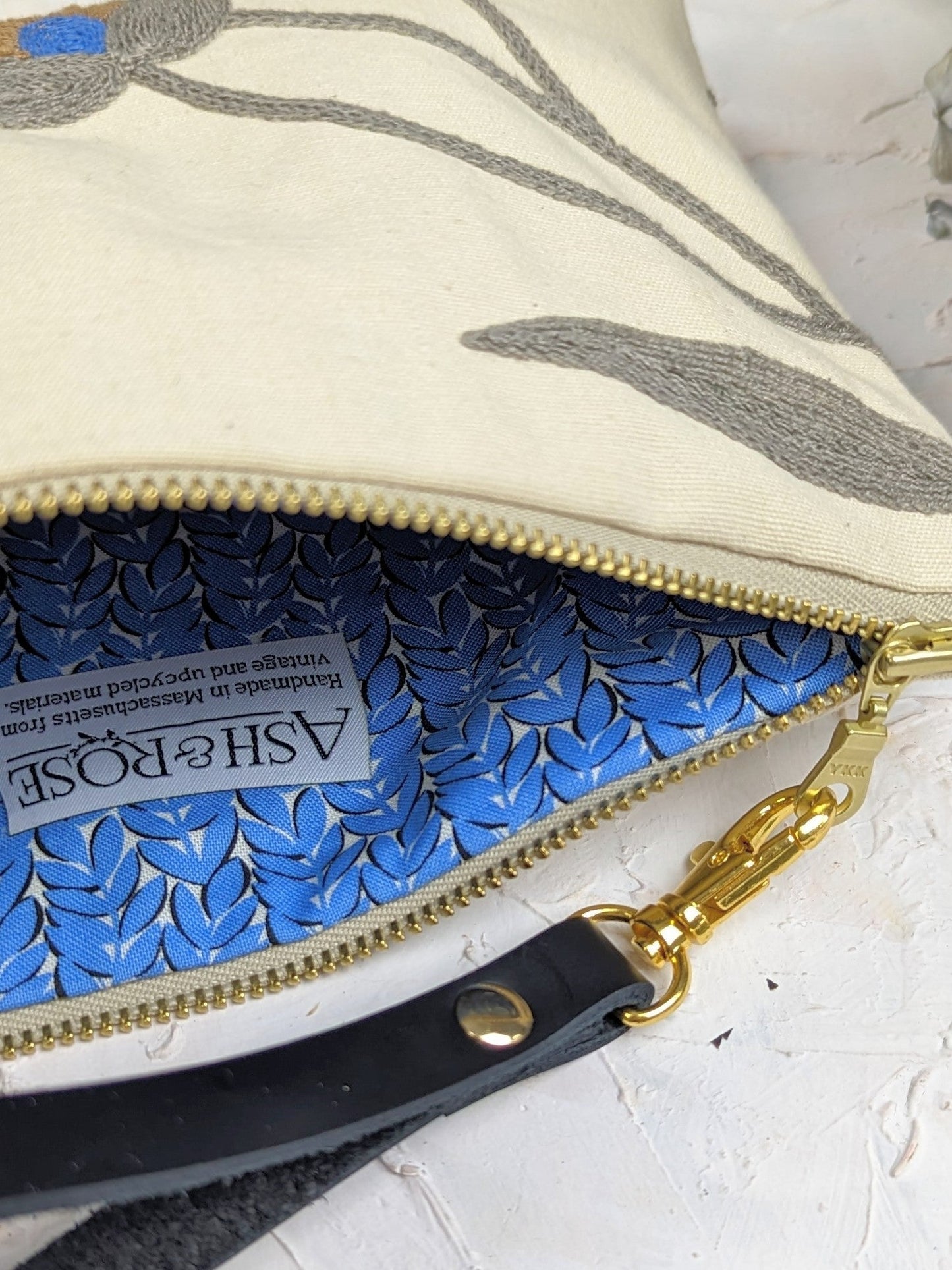 Close-up of an open Blue Poppy Wristlet Purse by Ash & Rose, featuring a cream linen exterior, gold zipper, blue patterned lining with an Ash & Rose label, and a black leather strap with a gold clip.