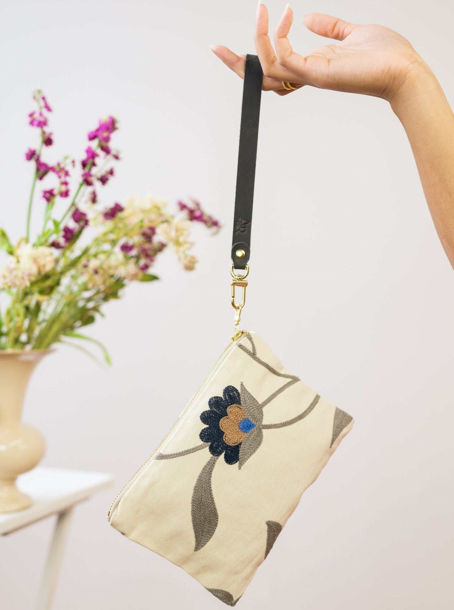 A hand holds the limited edition Ash & Rose Blue Poppy Wristlet Purse—a cream wristlet with a dark floral design and black strap—in front of a blurred vase of purple and white flowers on a table.