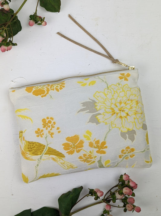 The Irene Song Bird Clutch by Ash & Rose features cream fabric with yellow birds and floral patterns, a gold zipper, and a beige wrist strap. It's displayed on a white surface with pink and green flower sprigs.