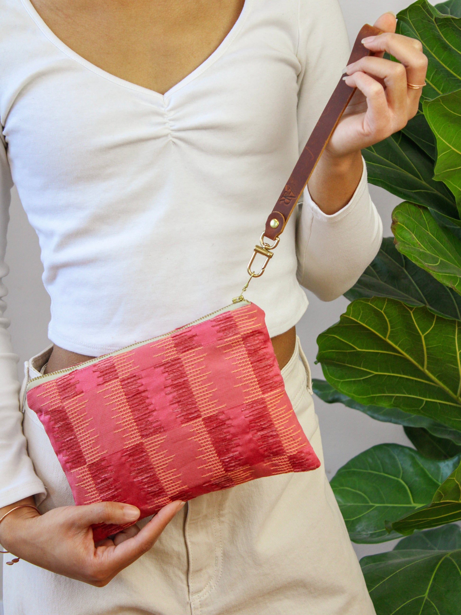 A person in a white long-sleeve top holds the Ash & Rose Shades of Apricot Wristlet Purse, a limited edition red and pink patterned clutch with a brown leather strap, against a backdrop of green fiddle-leaf fig leaves.