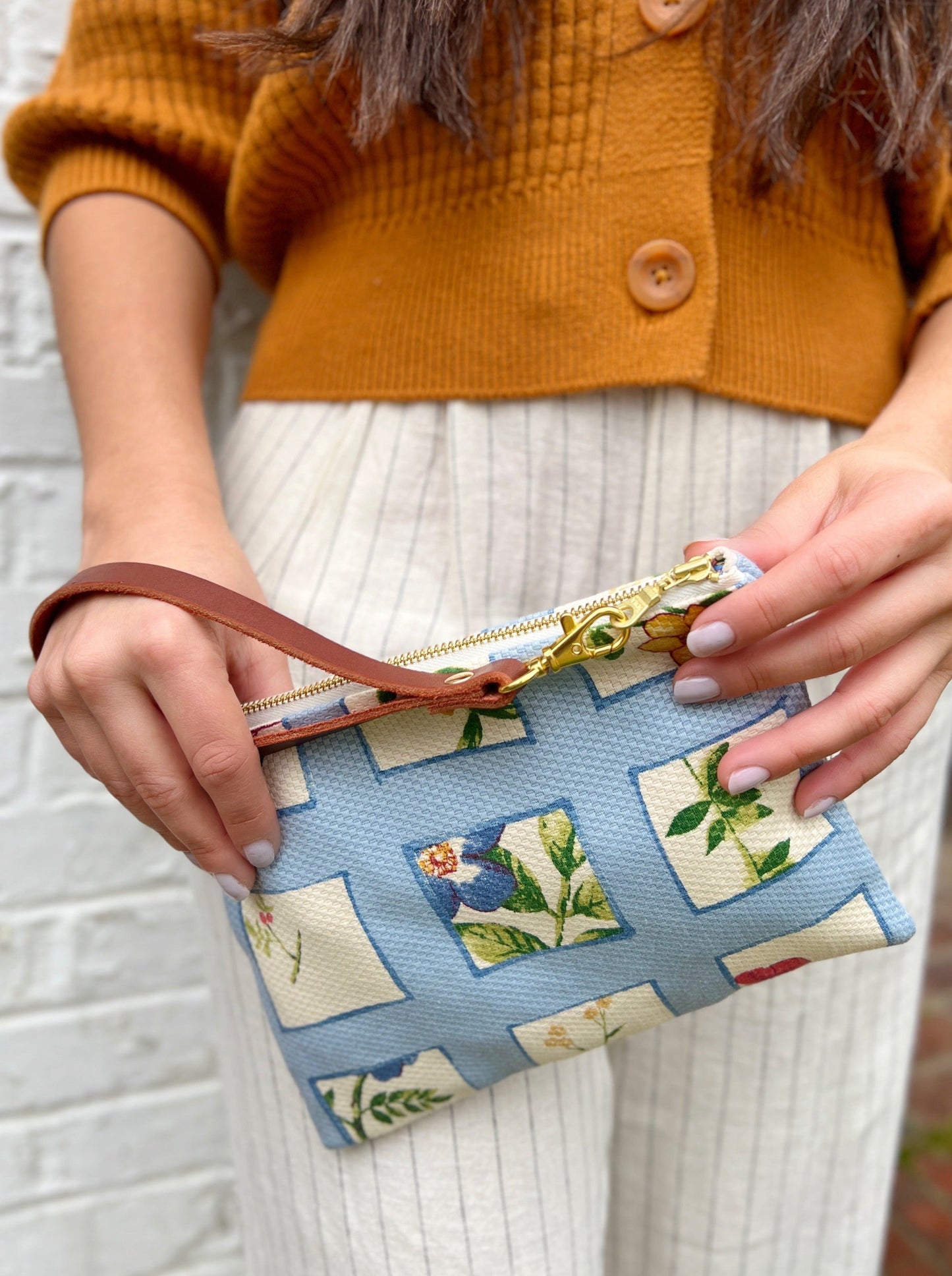 A person in a rust sweater and cream pinstriped pants holds the Ash & Rose Summer Meadow Wristlet Purse—a blue floral-patterned, handmade leather clutch with a brown strap and gold zipper.