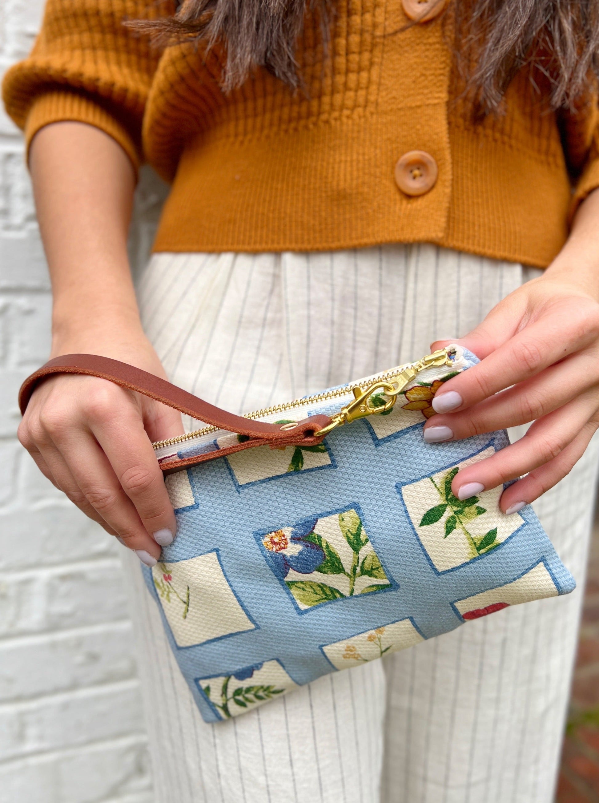 A person in a rust sweater and cream pinstriped pants holds the Ash & Rose Summer Meadow Wristlet Purse—a blue floral-patterned, handmade leather clutch with a brown strap and gold zipper.