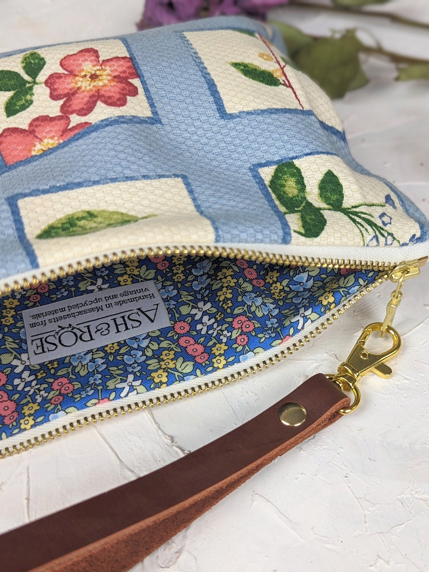 The Summer Meadow Wristlet Purse by Ash & Rose features a blue and white floral pattern, limited edition design, gold zipper, blue floral lining, brown handmade leather wrist strap with gold clasp, and an Ash & Rose label.
