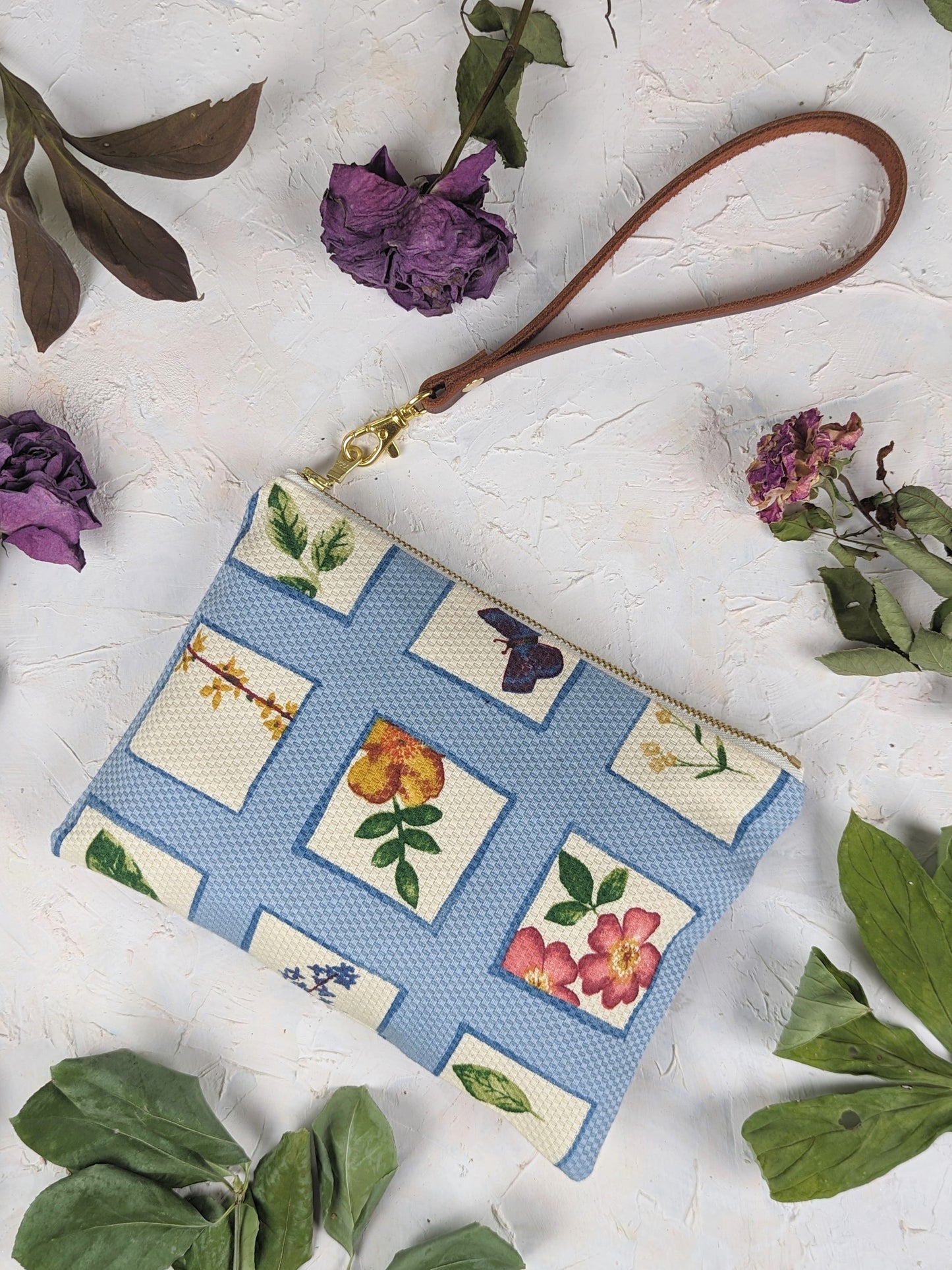 The Ash & Rose Summer Meadow Wristlet Purse is a blue limited edition clutch with a tan strap, zip closure, and grid pattern of colorful floral illustrations. Displayed on a light textured surface with dried purple flowers and green leaves.