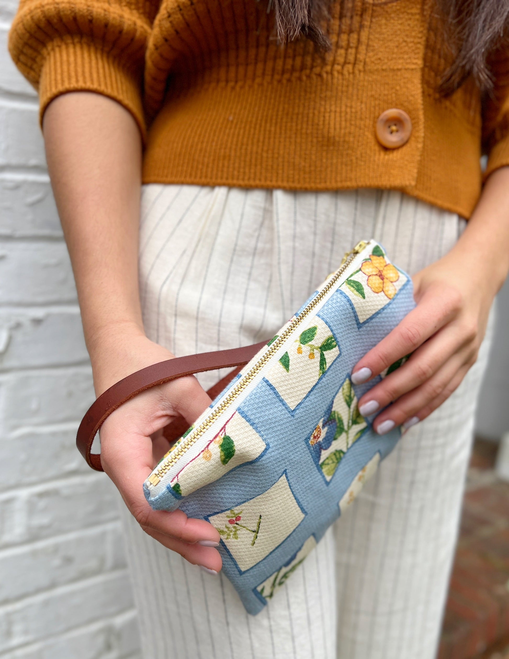 A person stands against a white brick wall, holding the Ash & Rose Summer Meadow Wristlet Purse—a vintage-style accessory with floral patchwork—while wearing a rust sweater and cream pinstriped pants.