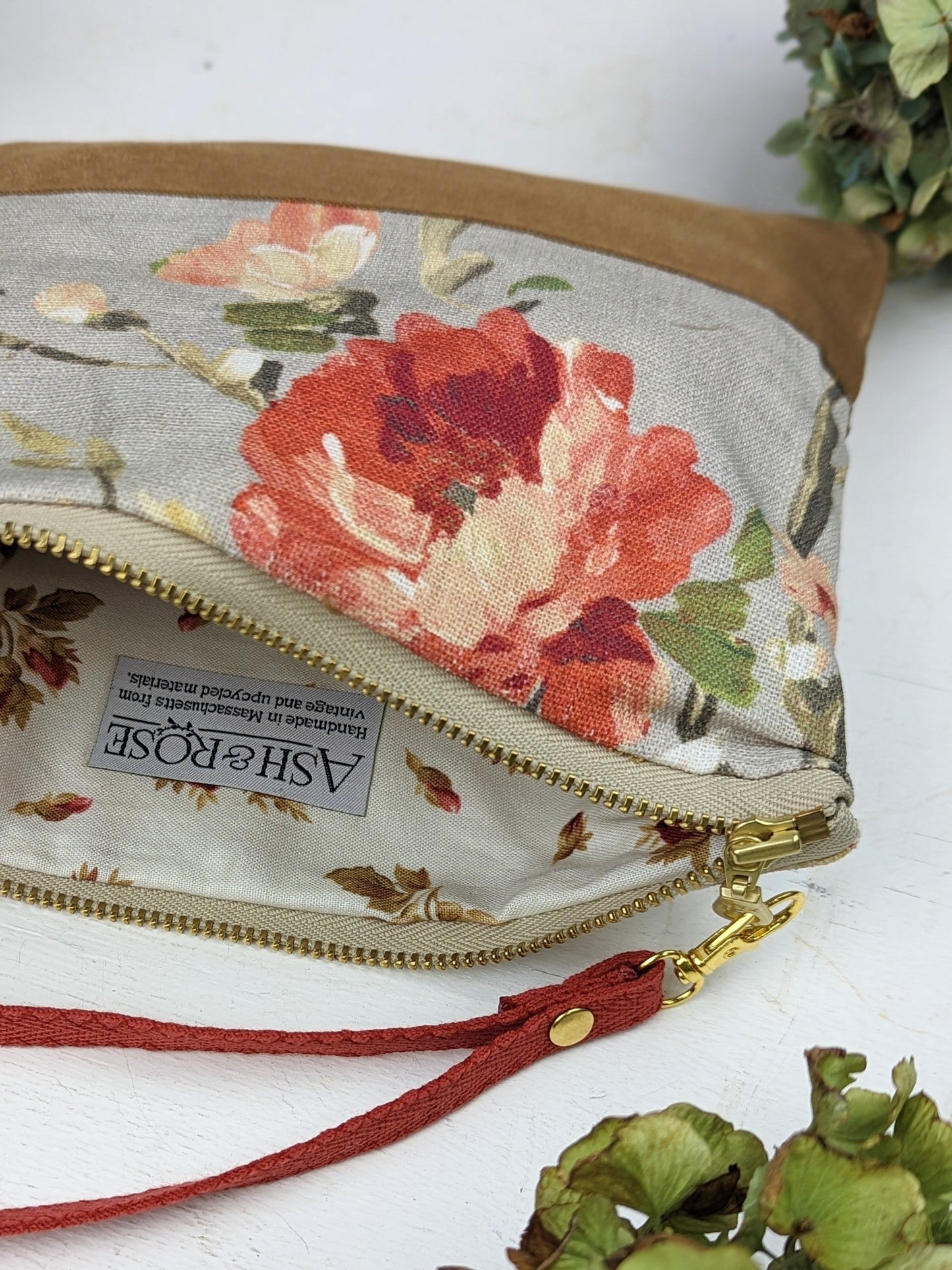 The Antique Roses Purse by Ash & Rose is a handmade cotton twill clutch with red, pink, and green floral patterns, a rose print wristlet strap, gold zipper, and matching floral lining. Green flowers surround the purse.