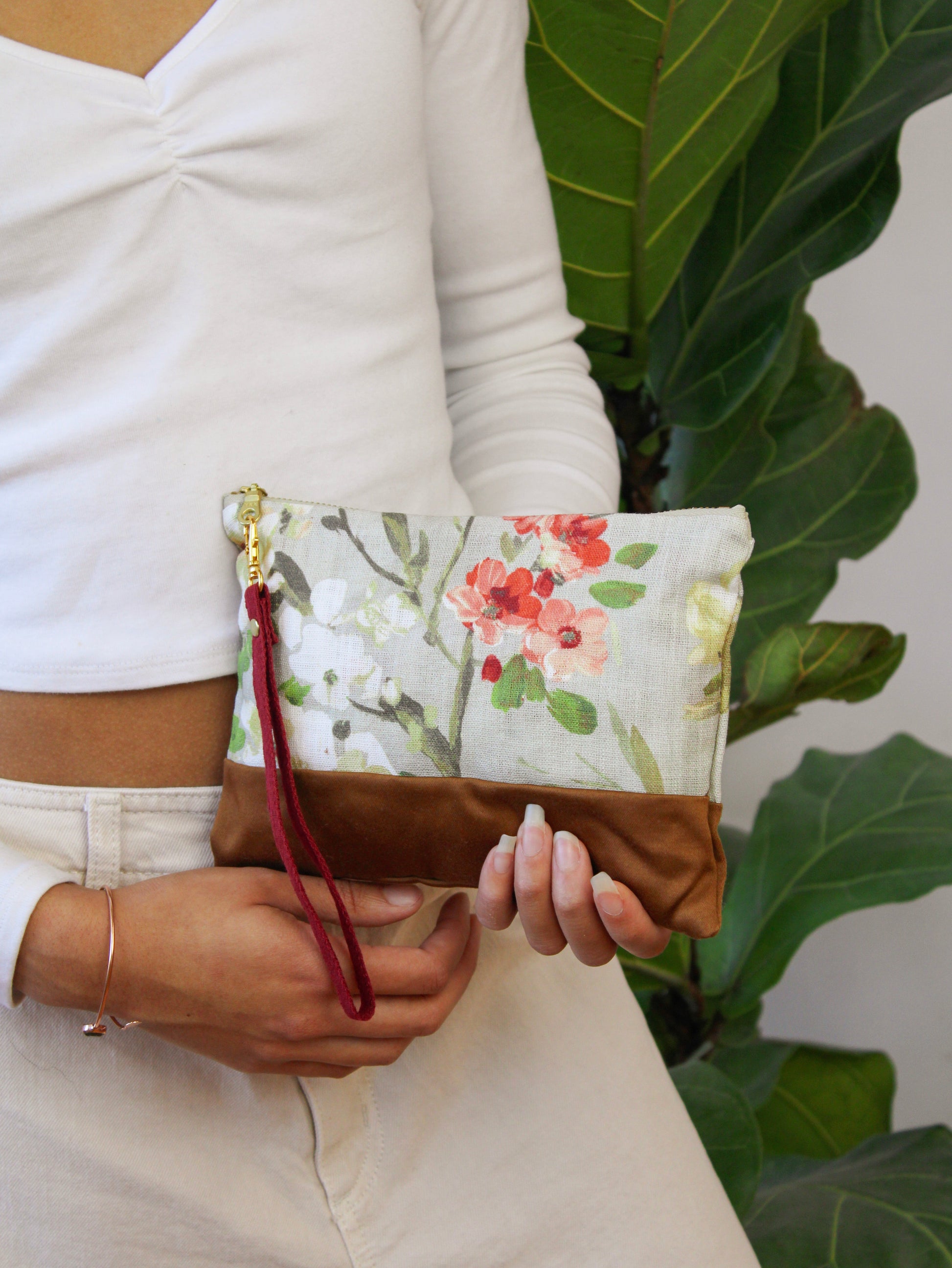 A person in a white long-sleeve top holds the Ash & Rose Antique Roses Purse, a handmade clutch with a floral pattern and brown base, standing before a large leafy plant.