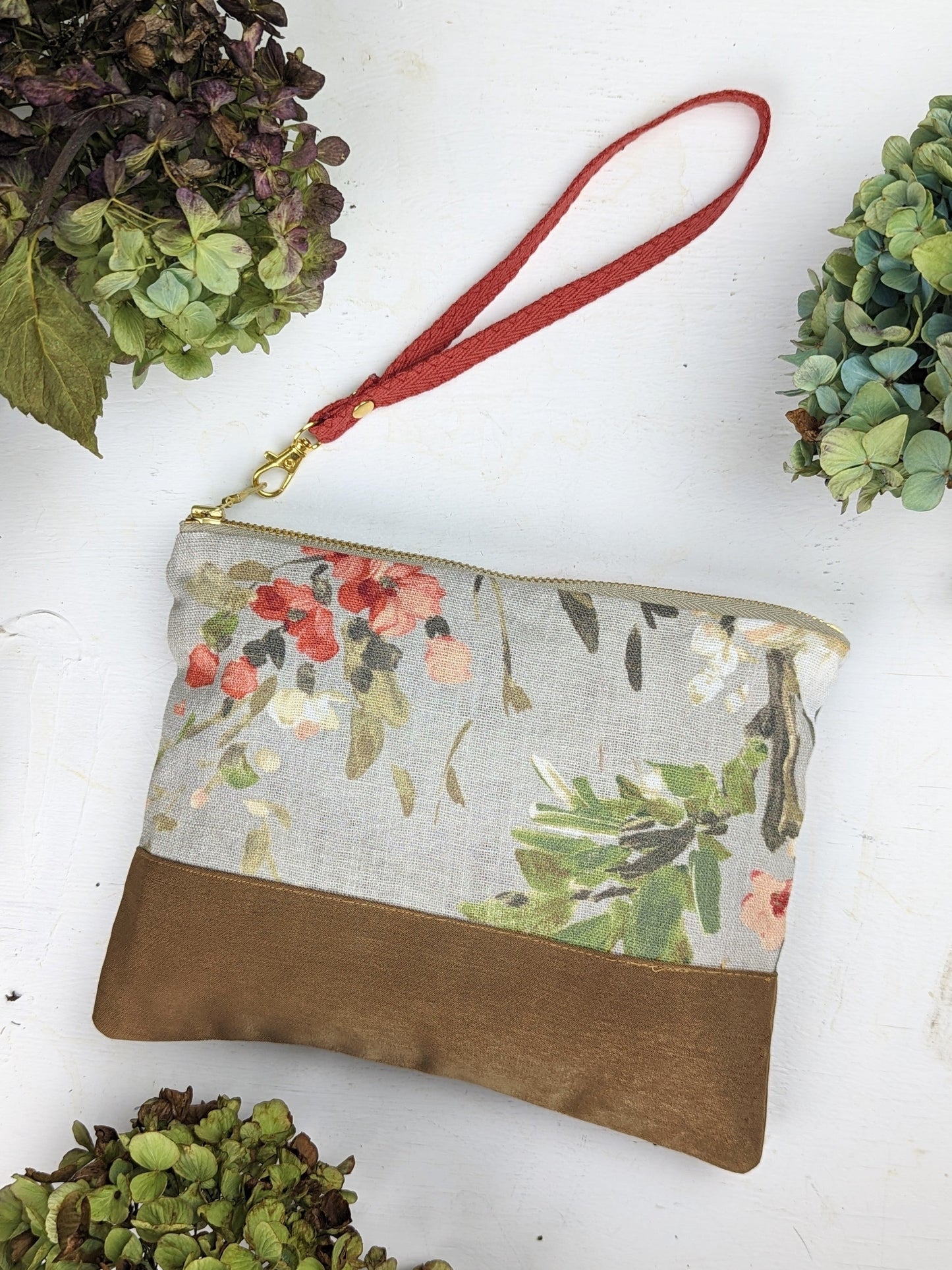The Ash & Rose Antique Roses Purse, featuring a rose print, brown bottom section, red wrist strap, and gold zipper, is shown on a white surface surrounded by green and purple hydrangeas.