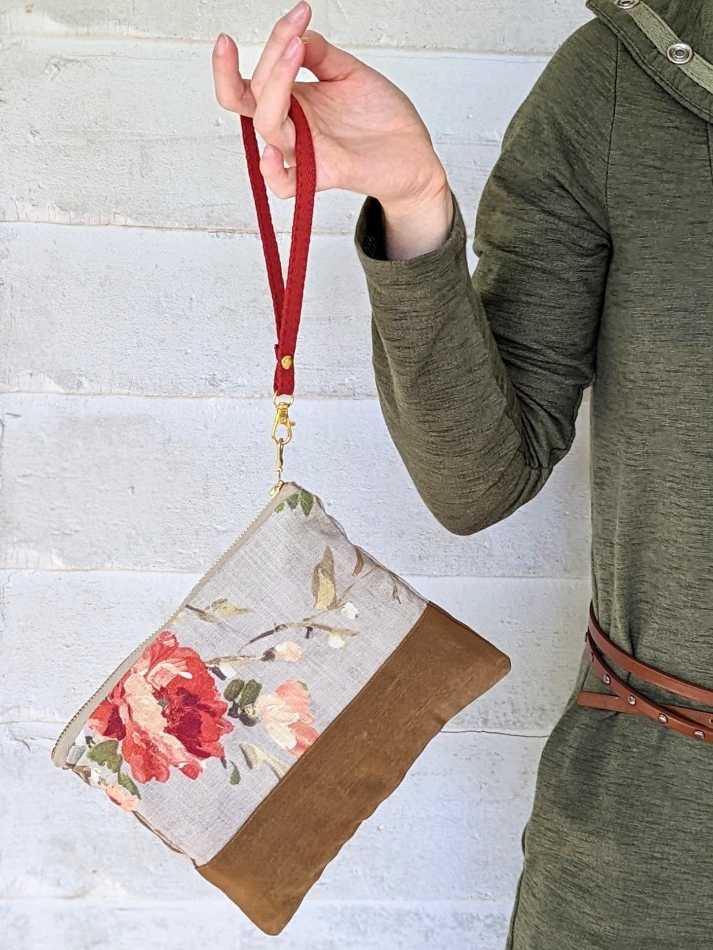 Wearing an olive green outfit, a person holds the Ash & Rose Antique Roses Purse—a handmade clutch with floral fabric, brown bottom, and red wrist strap—against a light-textured wall.