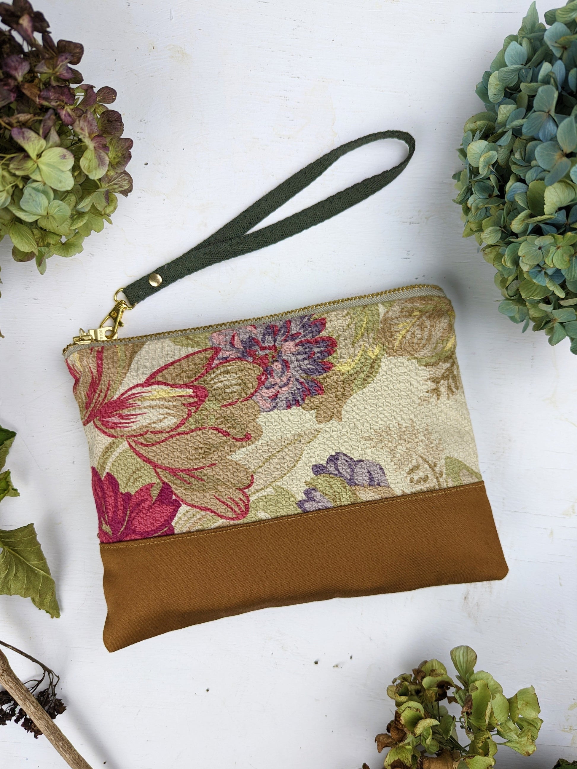The Ash & Rose Antique Roses Purse is a cotton twill wristlet with a tan bottom and green strap, shown on a white surface surrounded by green and purple hydrangeas.