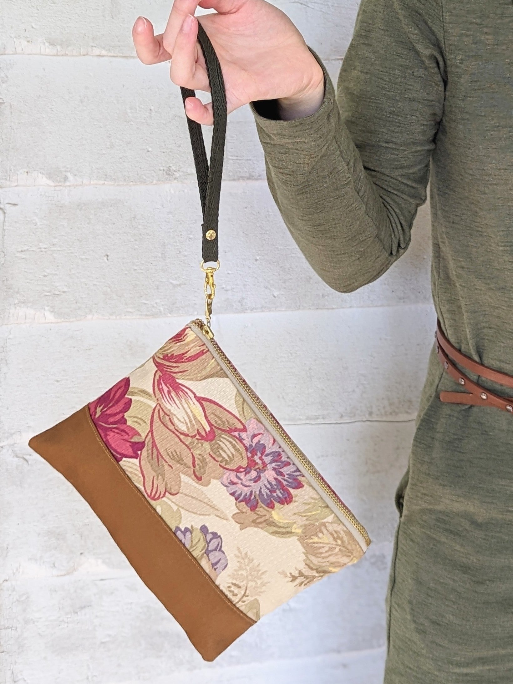 A person in a green dress holds the Ash & Rose Antique Roses Purse—a handmade wristlet clutch with a brown bottom and cream fabric featuring a beautiful pink, purple, and beige rose print—against a white brick background.