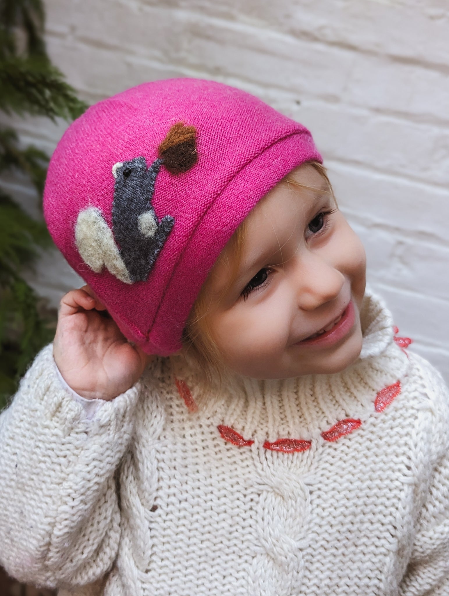 A smiling child wears the Ash & Rose Squirrel Cashmere Hat - Toddler, bright pink with a felt squirrel and acorn, paired with a cream sweater with pink accents. The child touches the hat while looking aside, against a white brick wall backdrop.