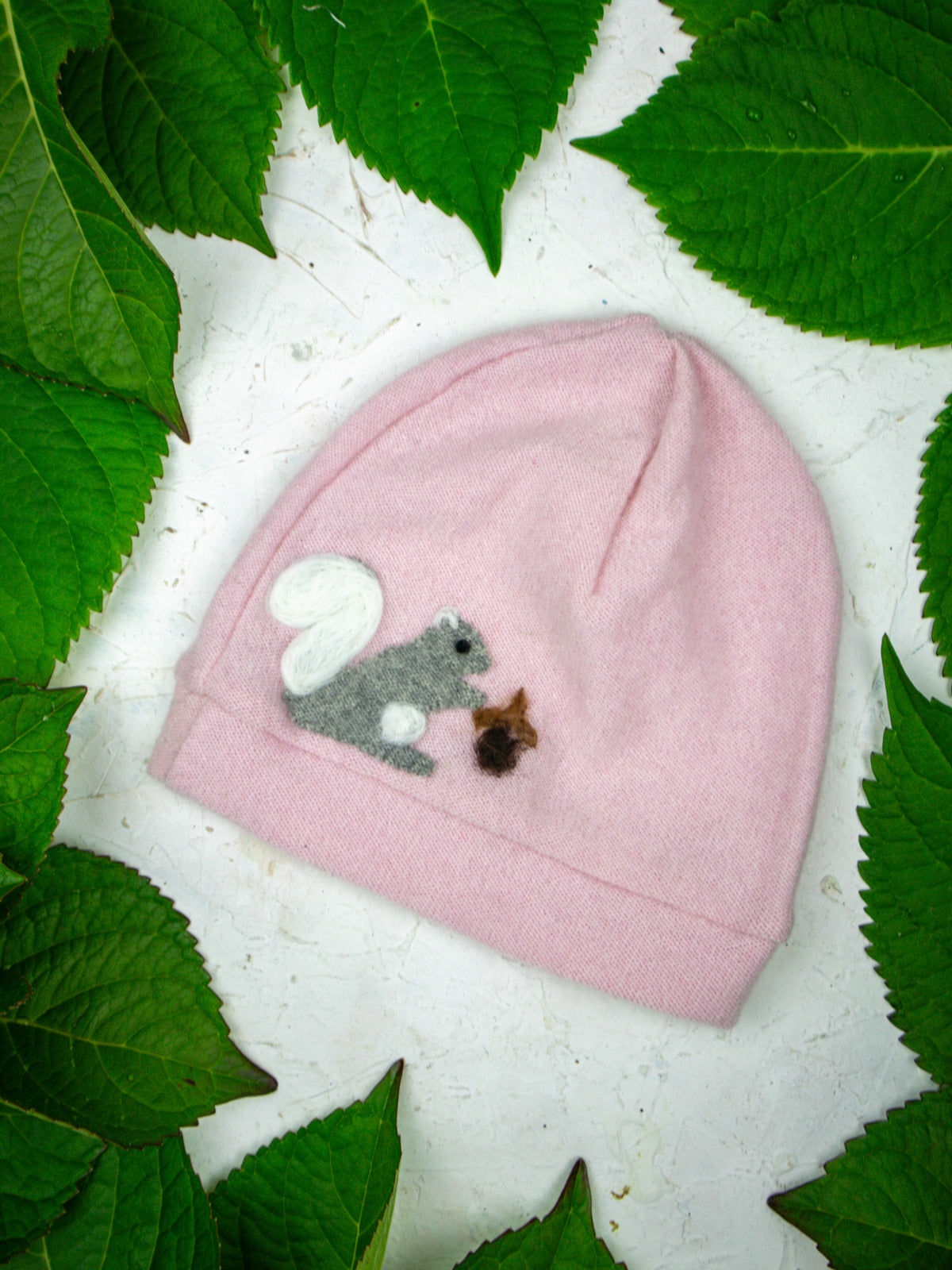 The Ash & Rose Squirrel Cashmere Hat - Toddler is a pink cashmere hat featuring a gray squirrel felt applique with an acorn, surrounded by green leaves on a white background.