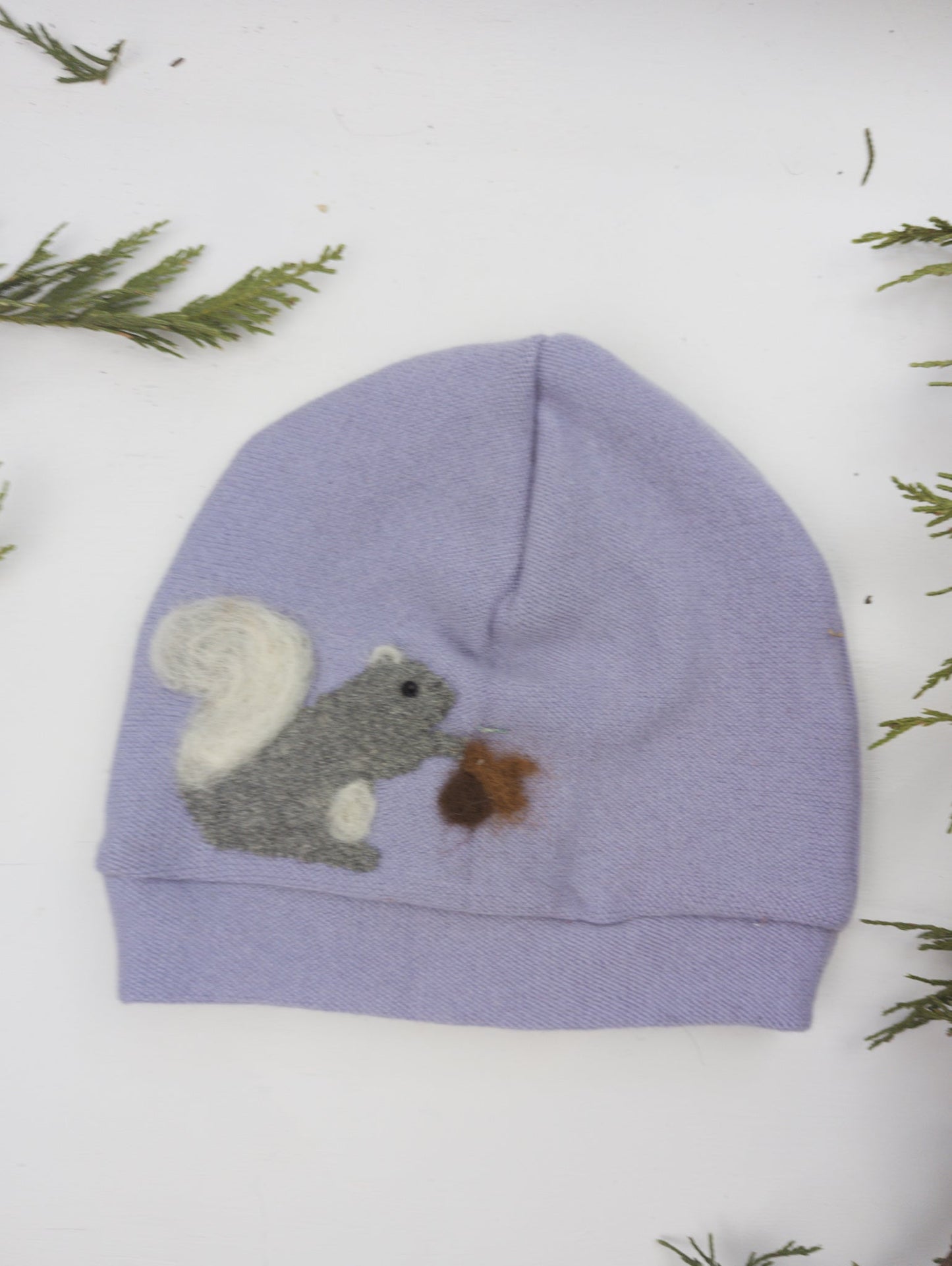 The Ash & Rose Squirrel Cashmere Hat - Toddler is a light purple recycled cashmere hat with a felted gray squirrel holding a brown acorn, surrounded by green foliage on a white background.