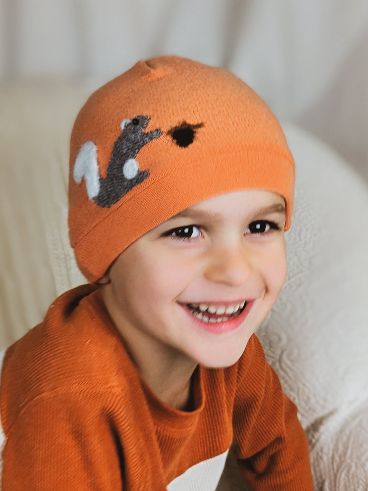A happy child wears the Ash & Rose Squirrel Cashmere Hat – Child, a handmade orange hat featuring a gray squirrel and acorn design, paired with an orange sweater and seated on a light-colored textured surface.