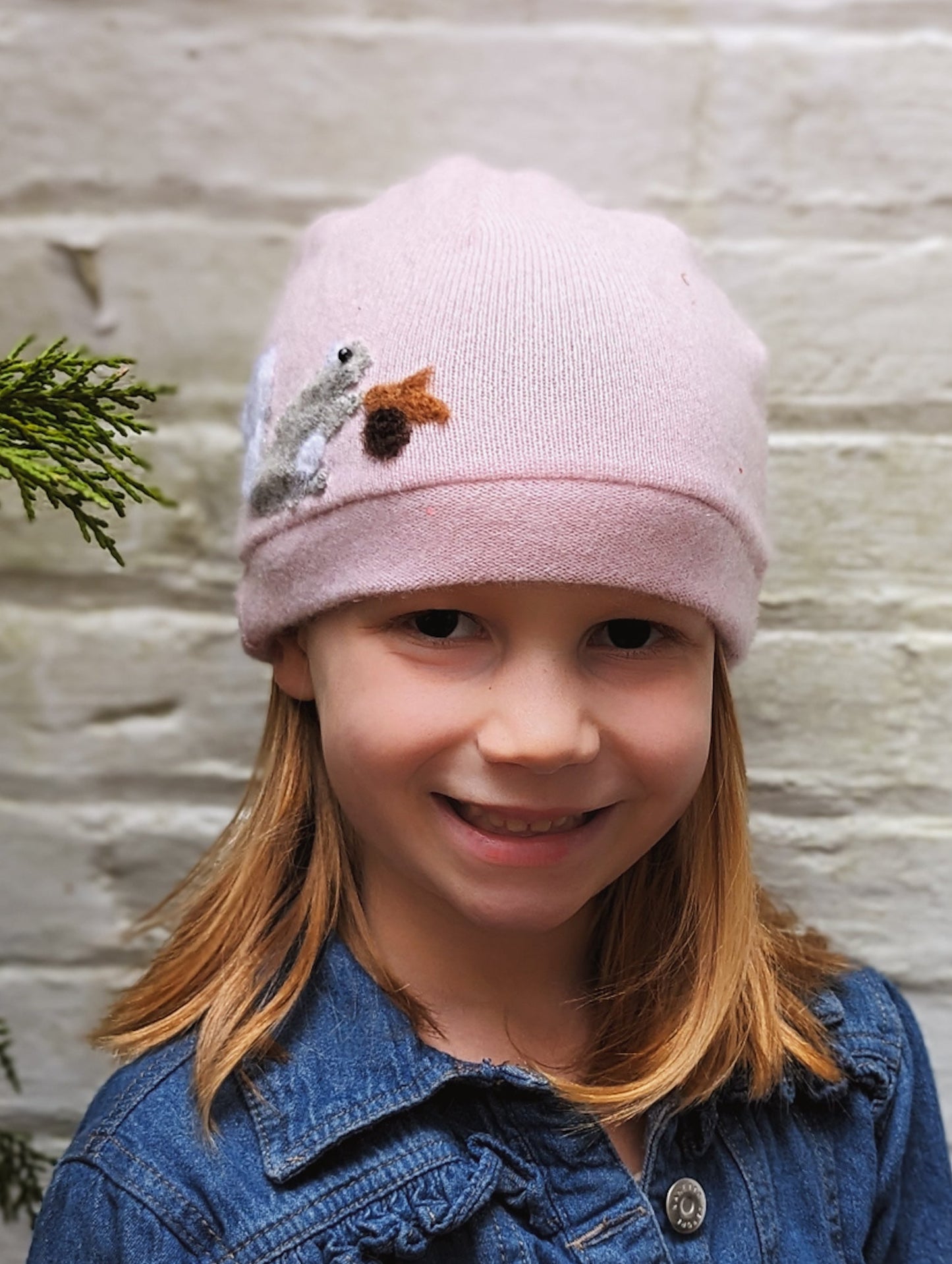 A young girl with straight blonde hair smiles at the camera, wearing the Ash & Rose Squirrel Cashmere Hat - Child, which is light pink with an embroidered squirrel on the front. She also wears a blue denim jacket. A green branch appears at the edge of the image.
