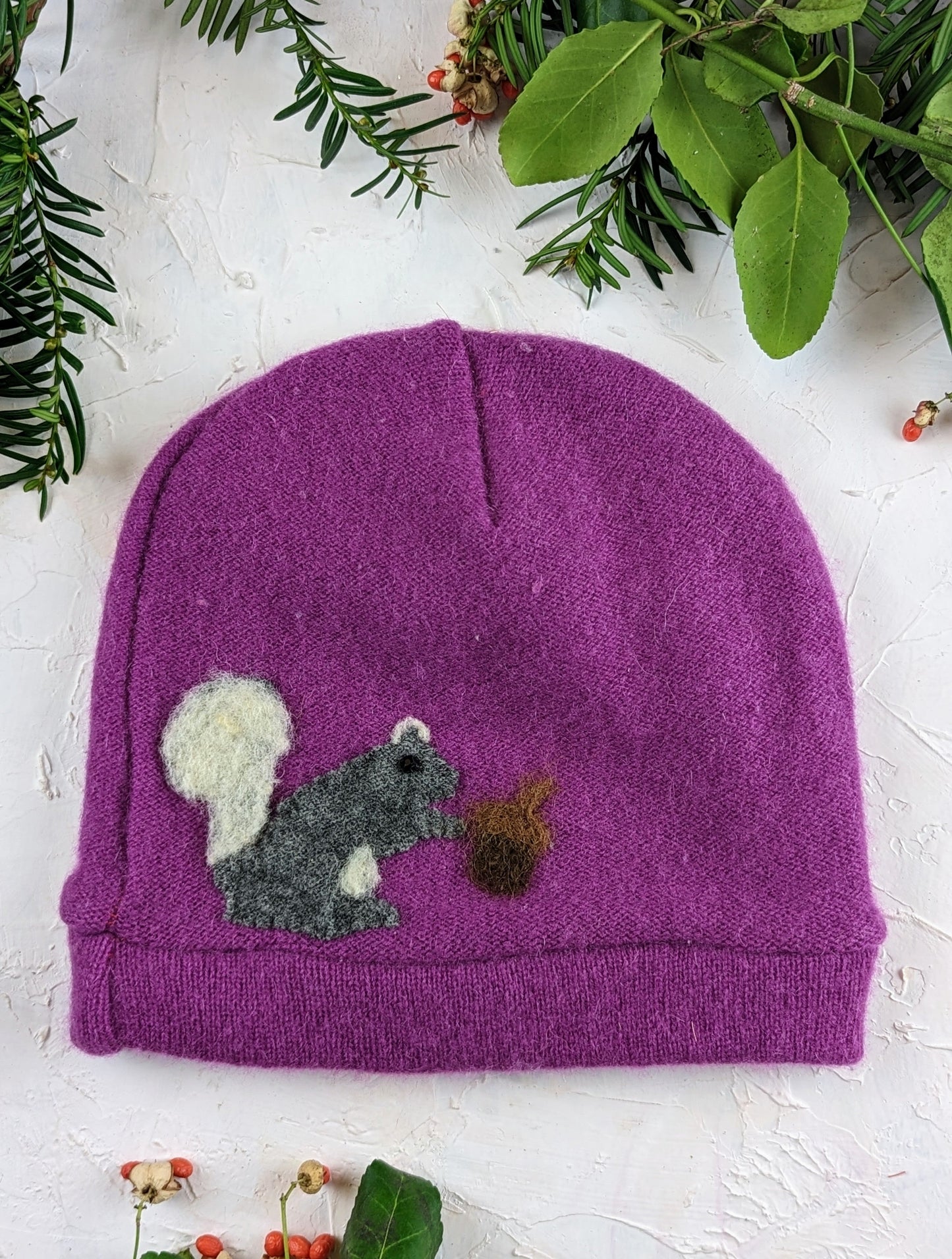 The Ash & Rose Squirrel Cashmere Hat - Child is a purple recycled cashmere hat with a felt gray squirrel holding an acorn, surrounded by green leaves and red berries, displayed on a white textured background.