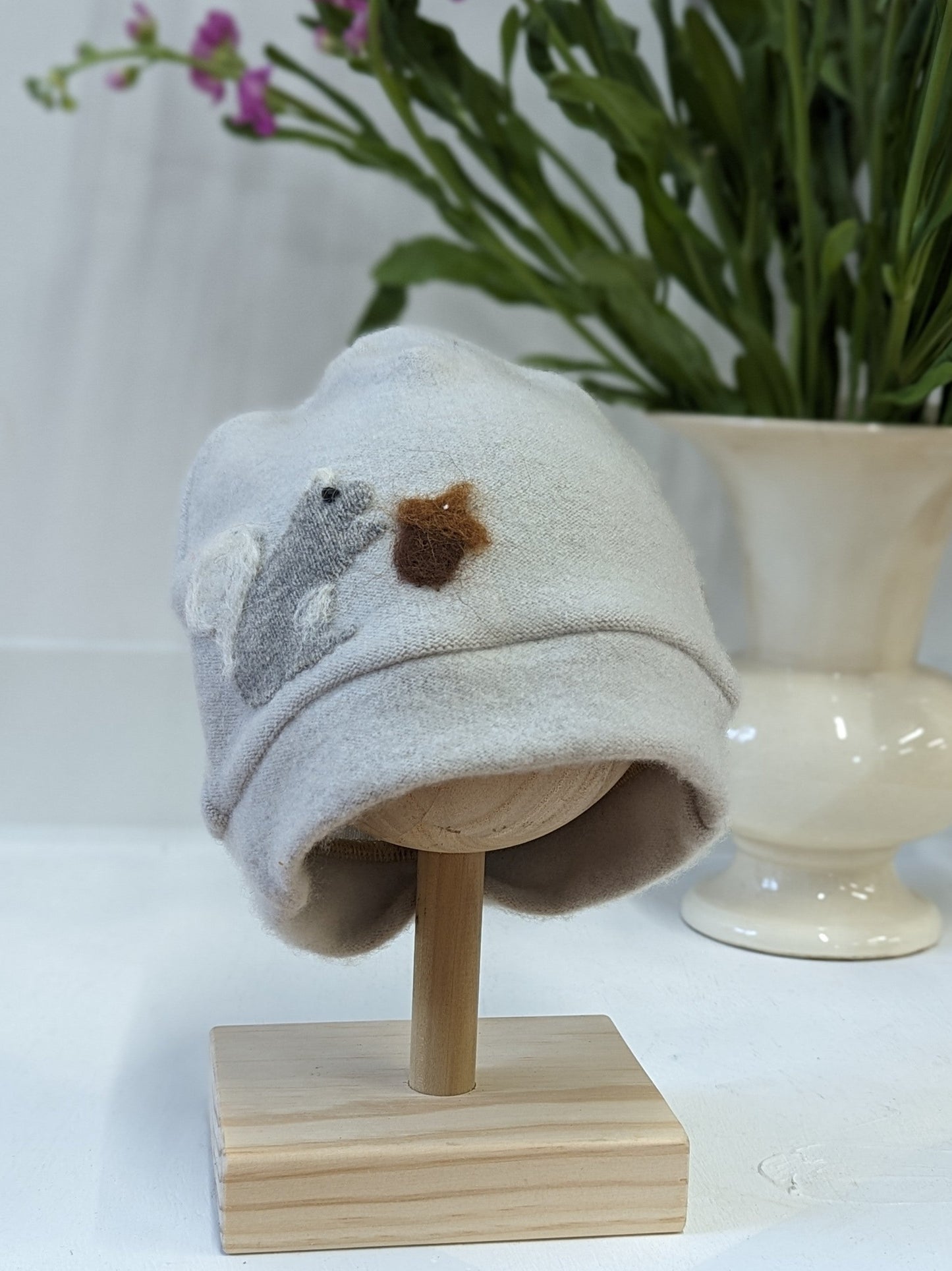 The Ash & Rose Squirrel Cashmere Hat - Child is a soft, light gray handmade kids’ hat from upcycled cashmere, featuring a felt gray squirrel eating brown cheese. Shown on a wooden stand with a white vase and green leafy plants behind.