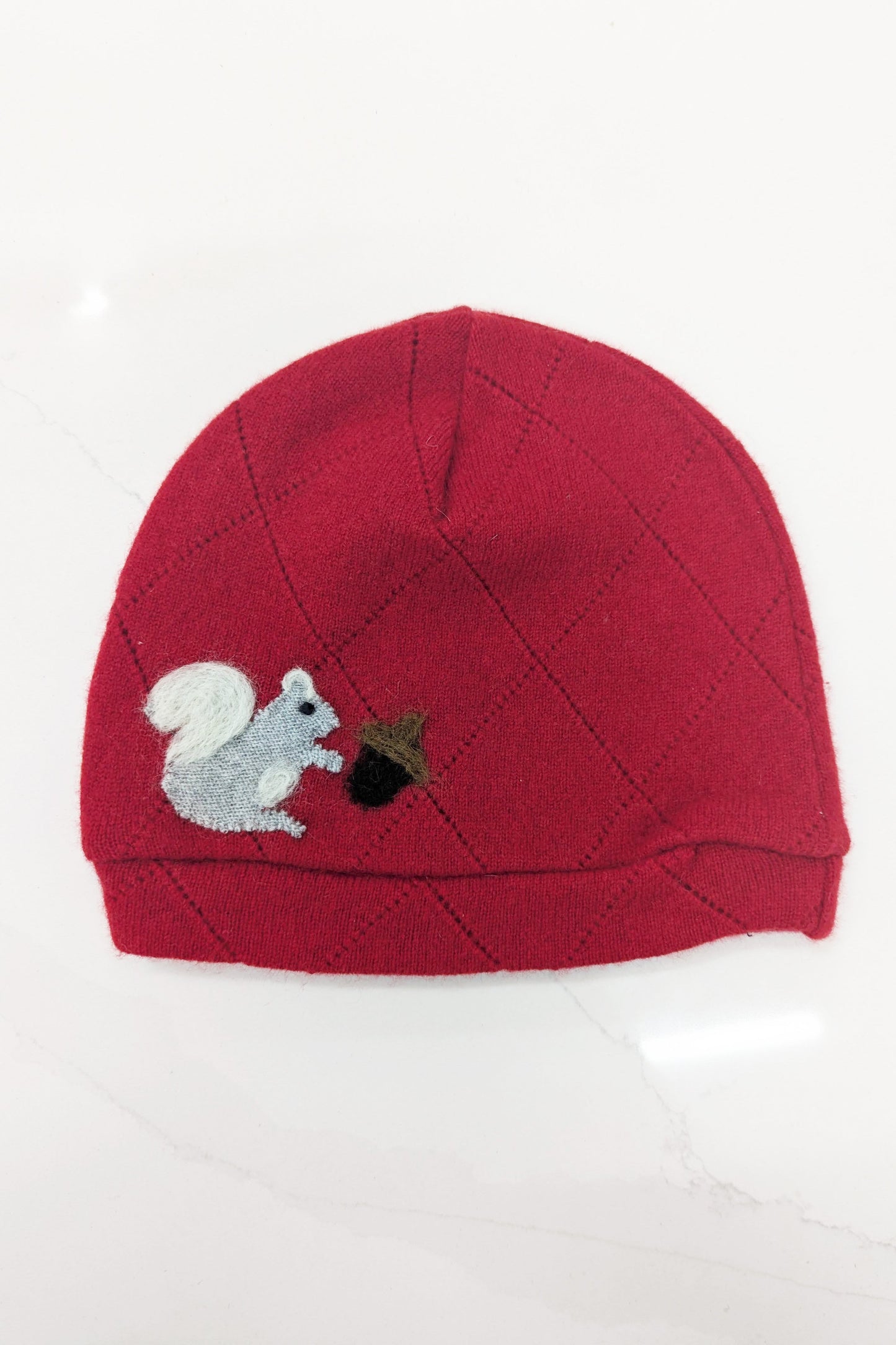 The Ash & Rose Squirrel Cashmere Hat - Child is a red upcycled cashmere knit beanie with a stitched pattern, featuring an embroidered gray squirrel holding a brown acorn on the front.