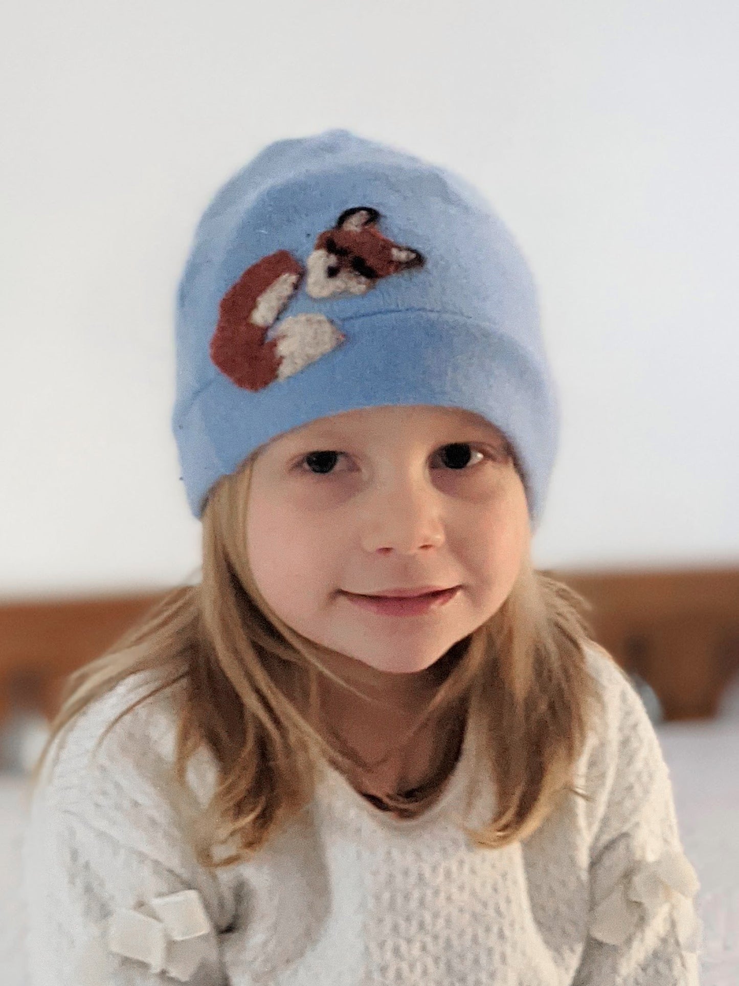 A young child with long blonde hair smiles softly in the Ash & Rose Sleeping Fox Cashmere Hat – Child, a handmade light blue kids hat featuring a fox design, paired with a white textured sweater against a blurred light background.
